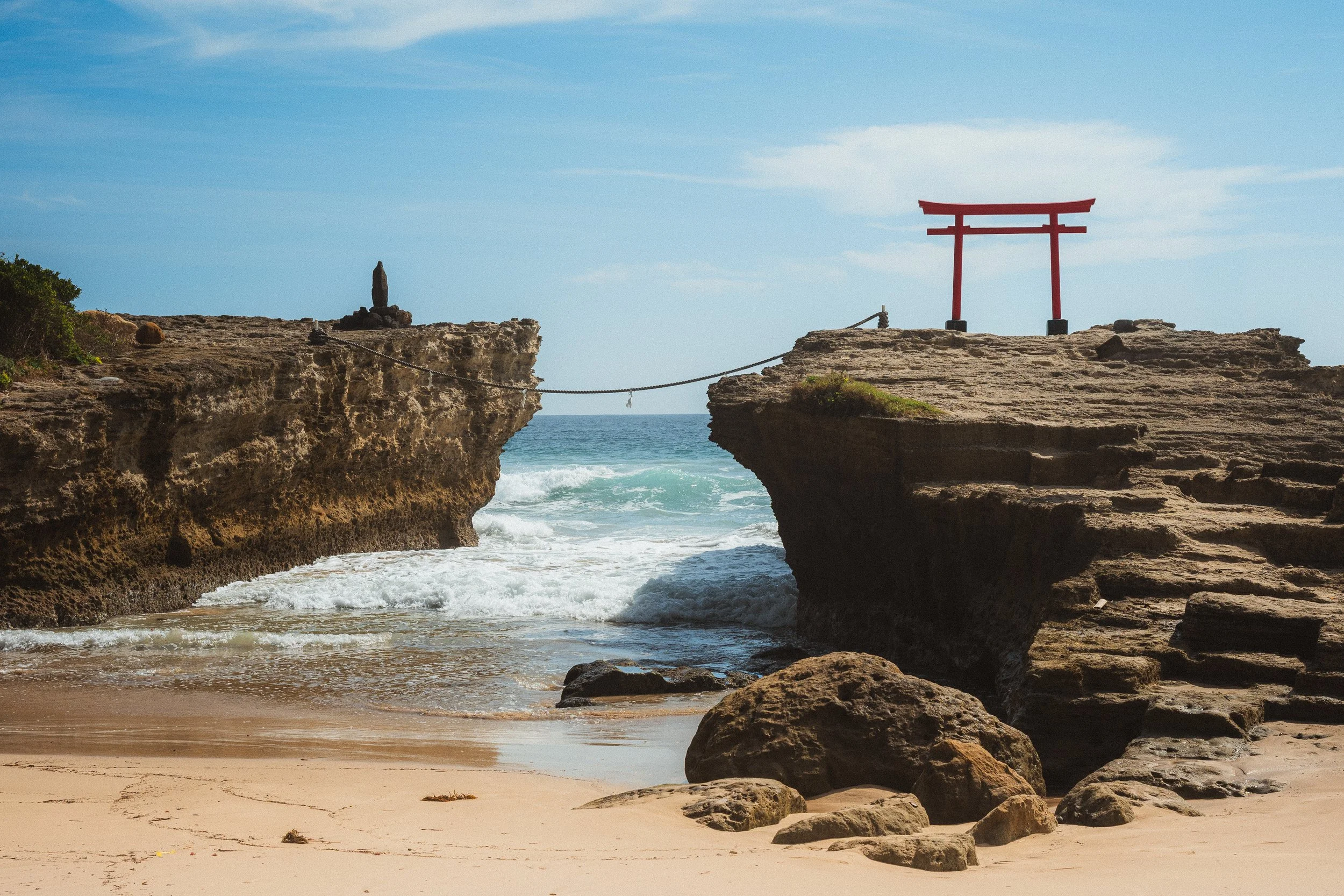 A beach scene with rocks and a cliff in the foreground, ocean waves, and a bright sky with some clouds. There is a red torii gate on the right rock formation and a small stone statue on the left cliff.