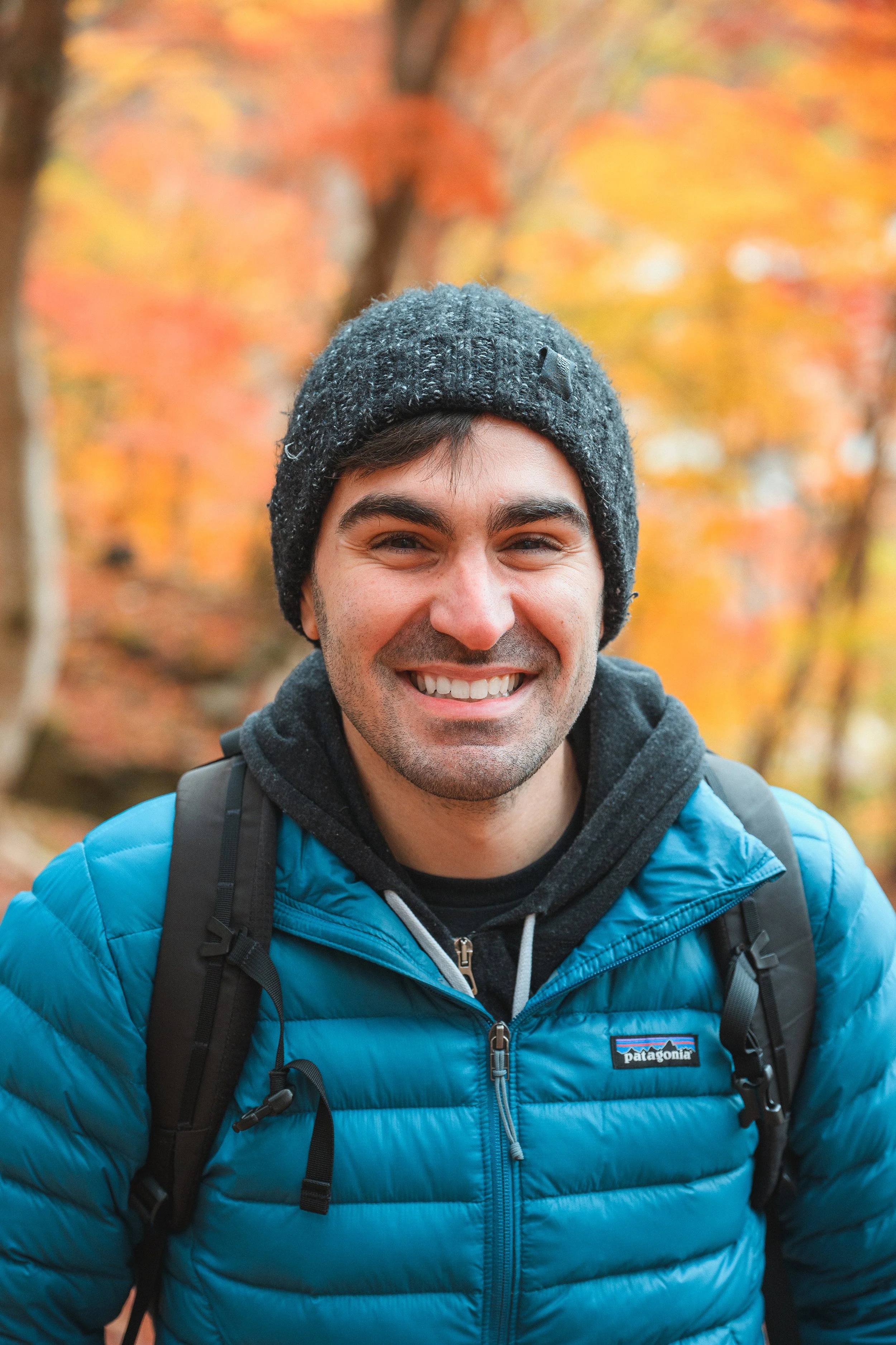 A smiling man wearing a black beanie, a blue Patagonia puffer jacket, and a black backpack, outdoors during fall with orange and yellow leaves in the background.
