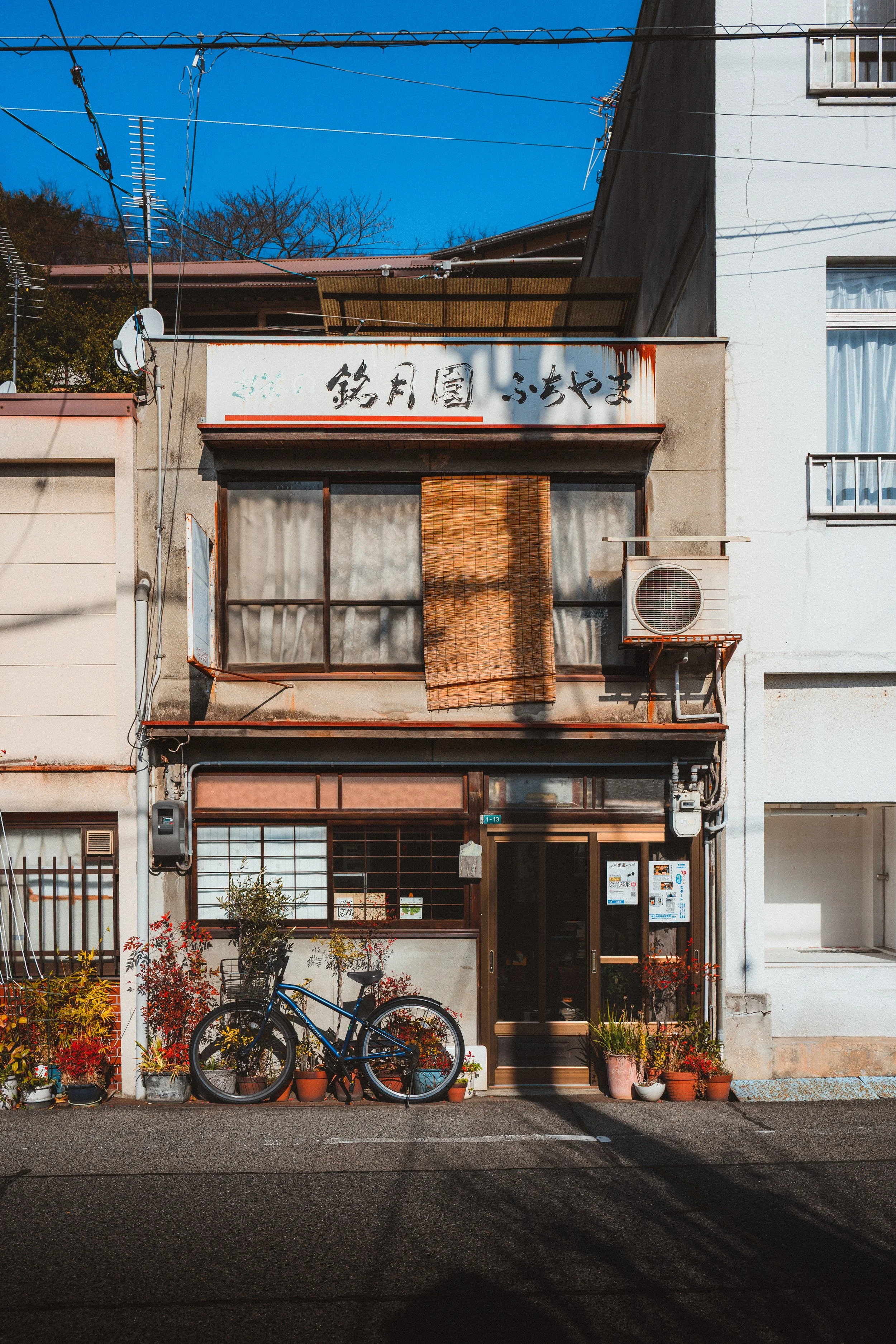 Front of a multi-story building with Japanese characters on a sign, surrounded by potted plants and flowers, two bicycles parked in front, and a blue sky overhead.