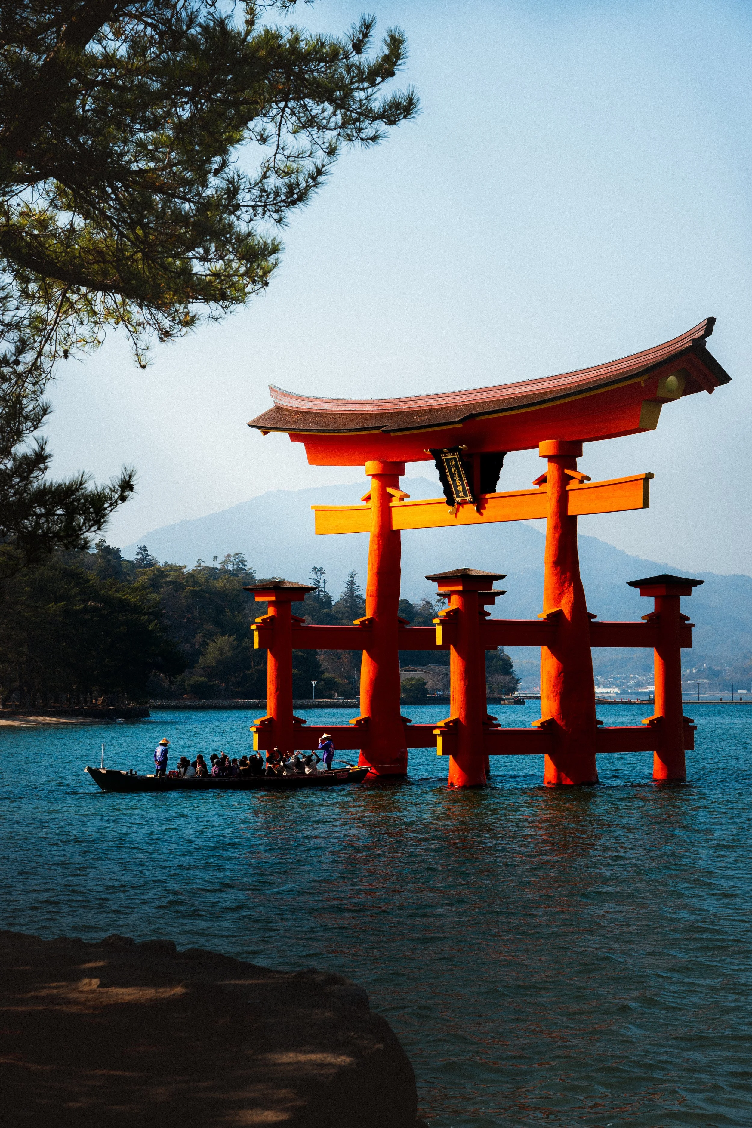 A large, red traditional Japanese torii gate standing in a body of water with mountains and trees in the background. A boat filled with people is near the torii gate, with some individuals wearing hats and life jackets.