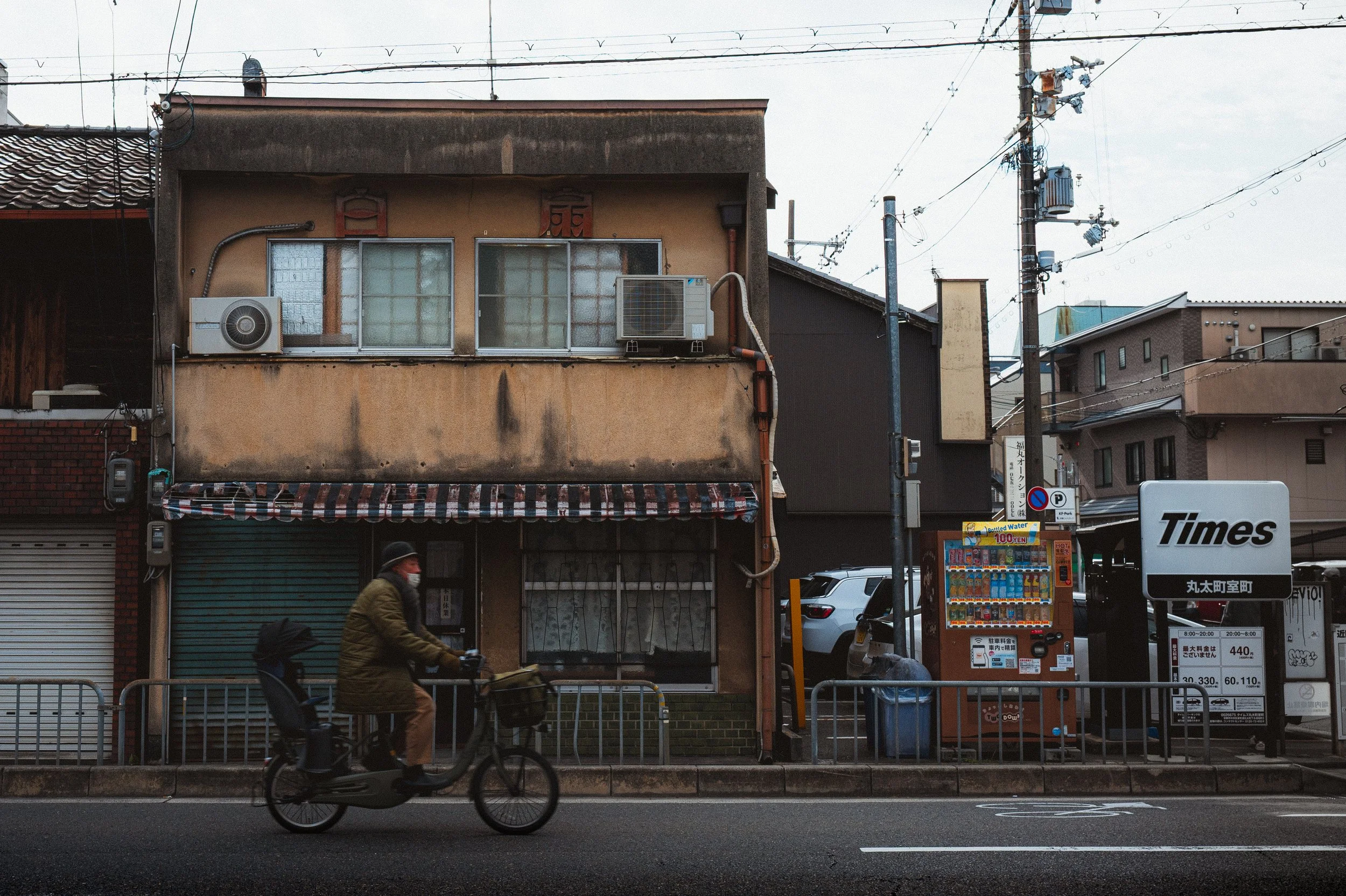 City street scene with a man riding an electric bicycle in front of a storefront with closed shutters, a person on a bicycle, and various buildings and utility poles.