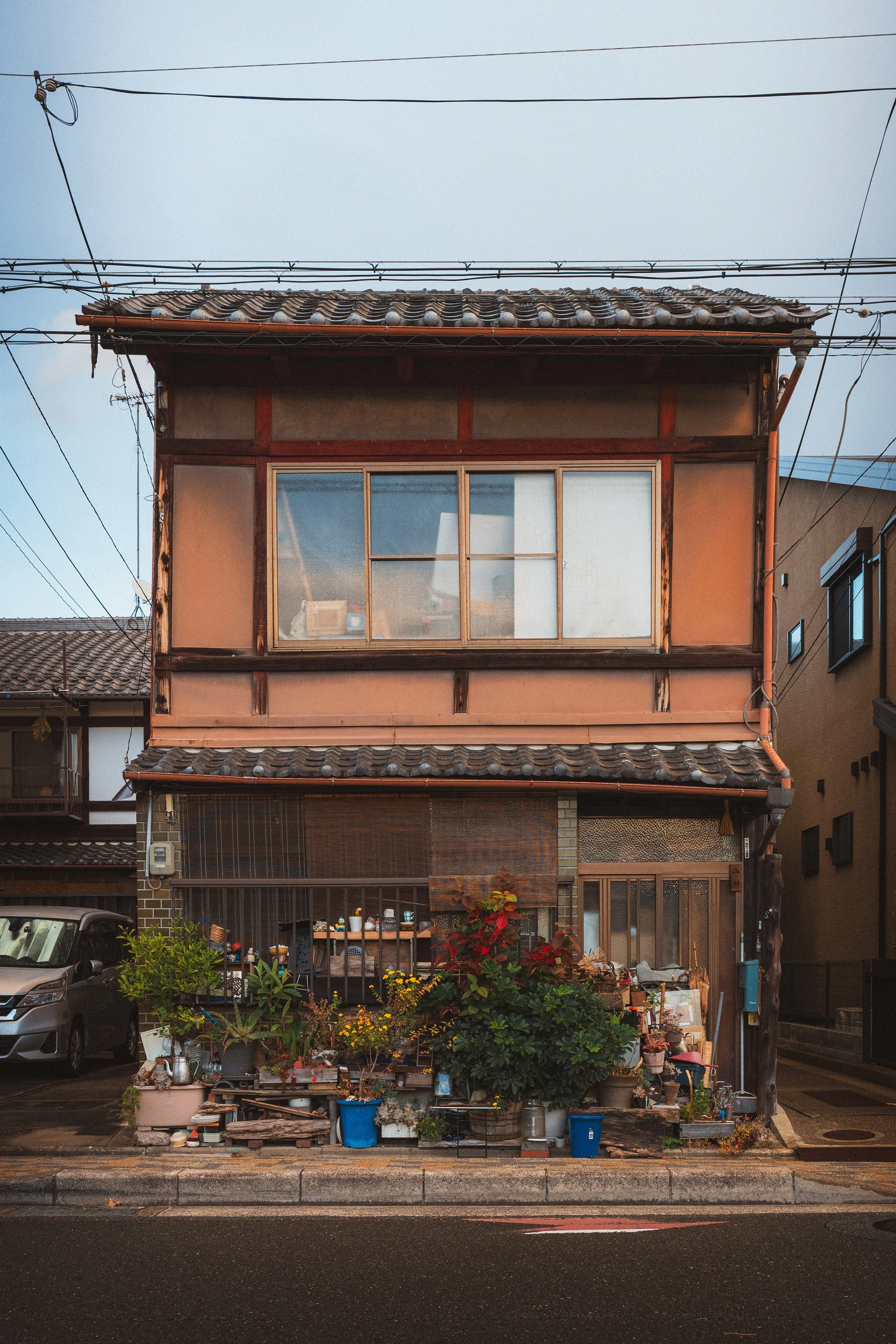 A multi-story building with a traditional tiled roof and large windows on the upper floor. The ground level features a garden with plants, flowers, and outdoor items, and a parked car is visible on the left. Overhead, electrical wires are strung acro