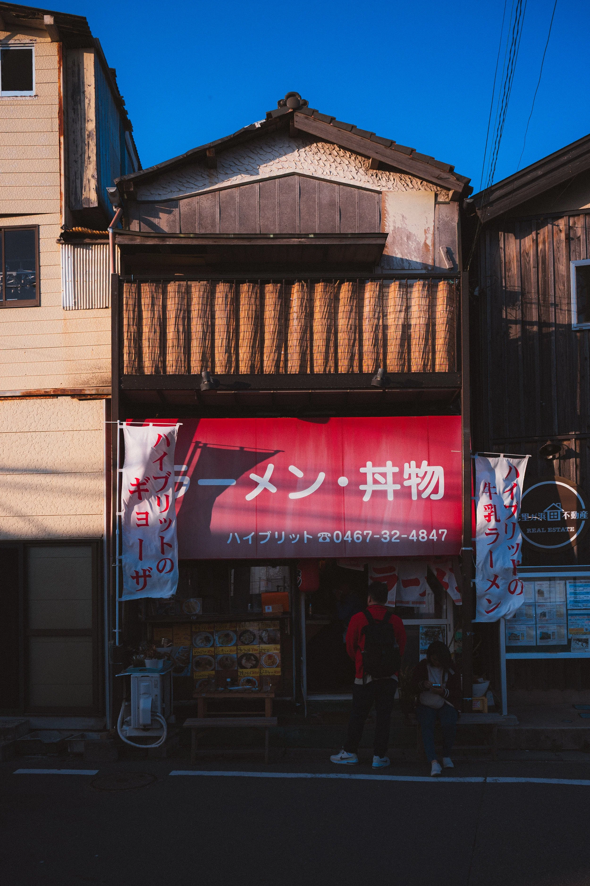 A street view of a Japanese restaurant with a red sign, white banners with red and blue writing, and an entrance with people standing outside. The building has a mix of wood and concrete exterior with a balcony.
