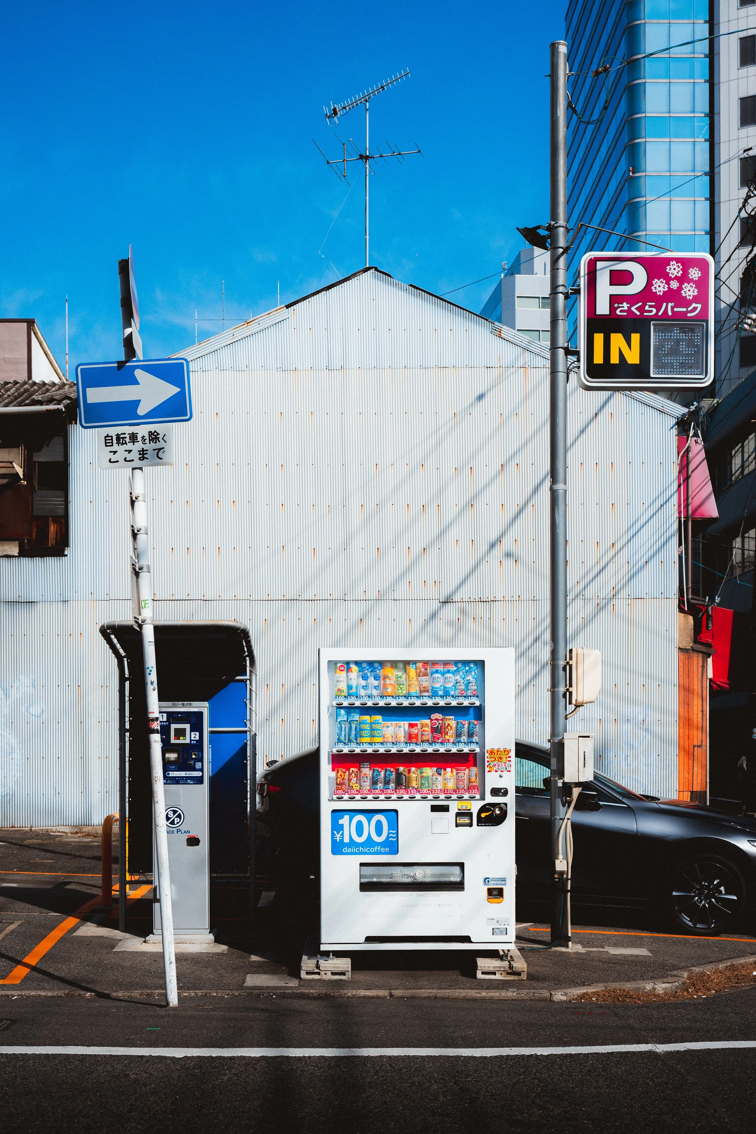 Japanese vending machine selling beverages, positioned under power lines, with a parking lot and a building with corrugated metal siding behind it. A blue arrow sign directs traffic to the right.