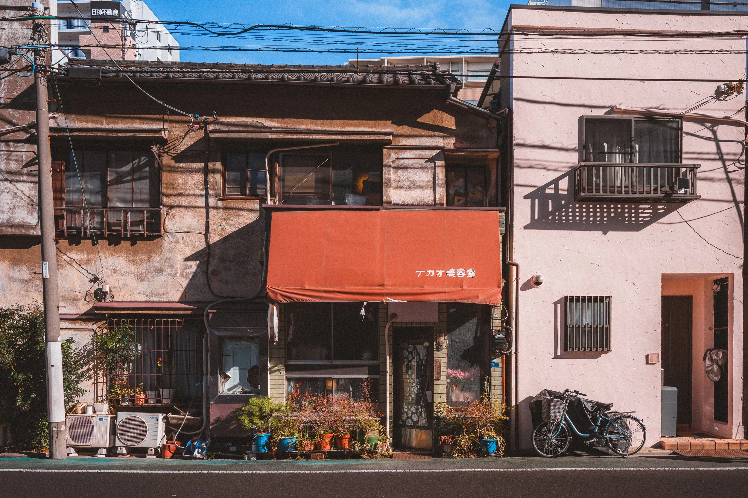 A street view of a building with a pink and brown facade, featuring balconies with railings, windows with bars, electric wires, a bicycle, potted plants, and an orange awning with Japanese characters. The sky is blue with some clouds.