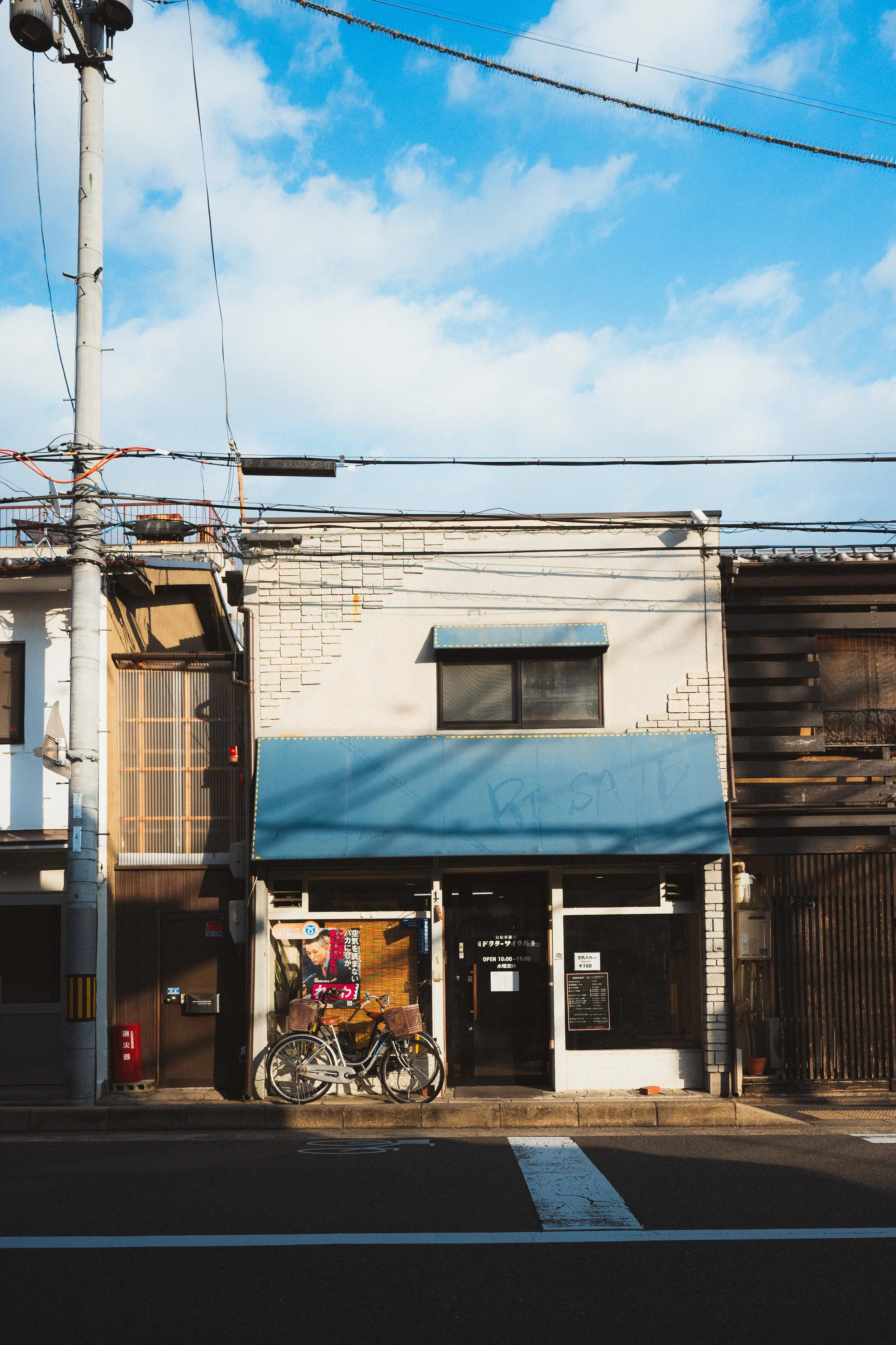 The image shows the front of a small building with a black door and a window, several bicycles parked in front, and a large blue sign with partially visible graffiti. There are electrical wires and a utility pole overhead, and a bright blue sky with 