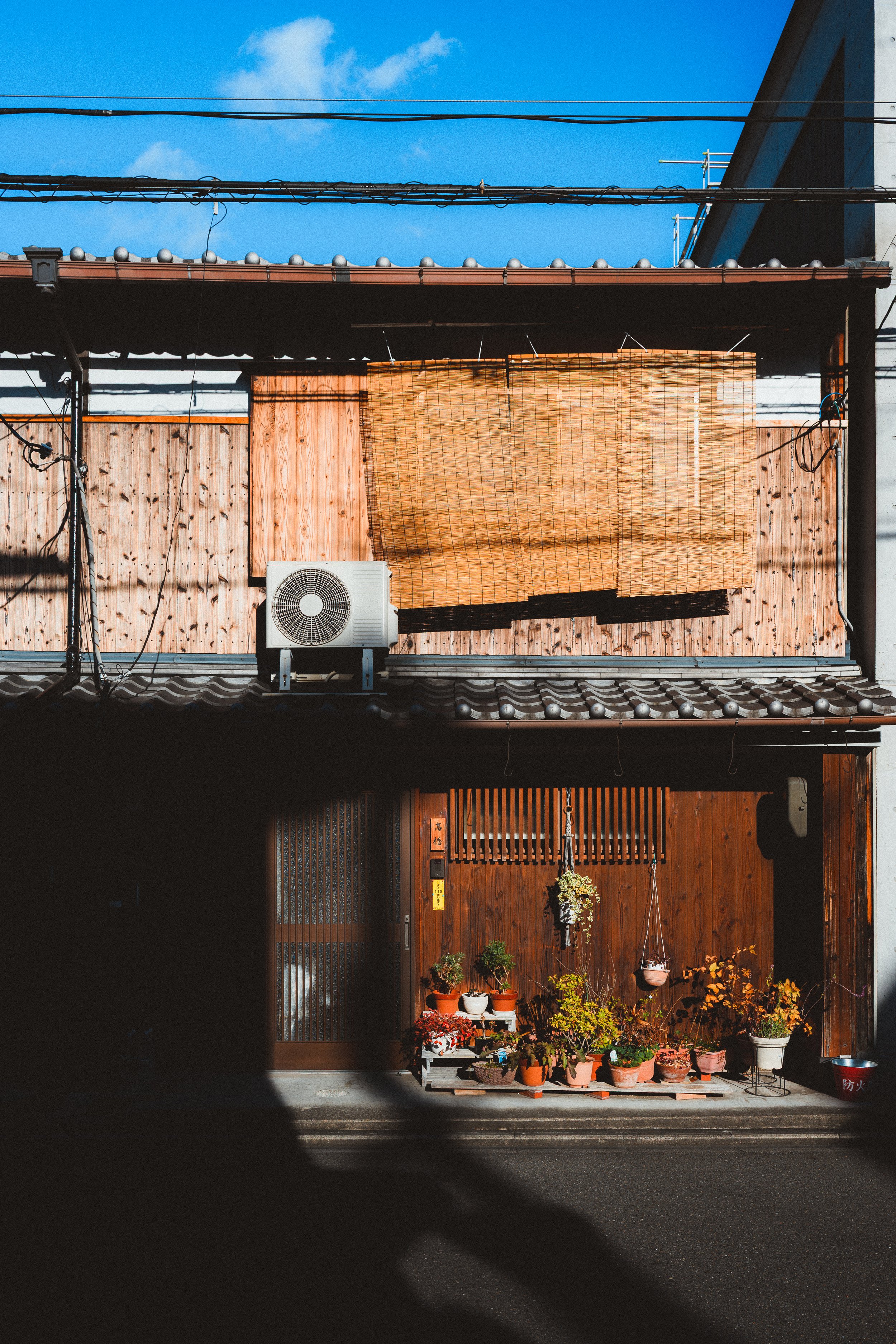Facade of a house with a wooden front, potted plants at the entrance, a bamboo screen, electrical wires overhead, and a blue sky with some clouds.