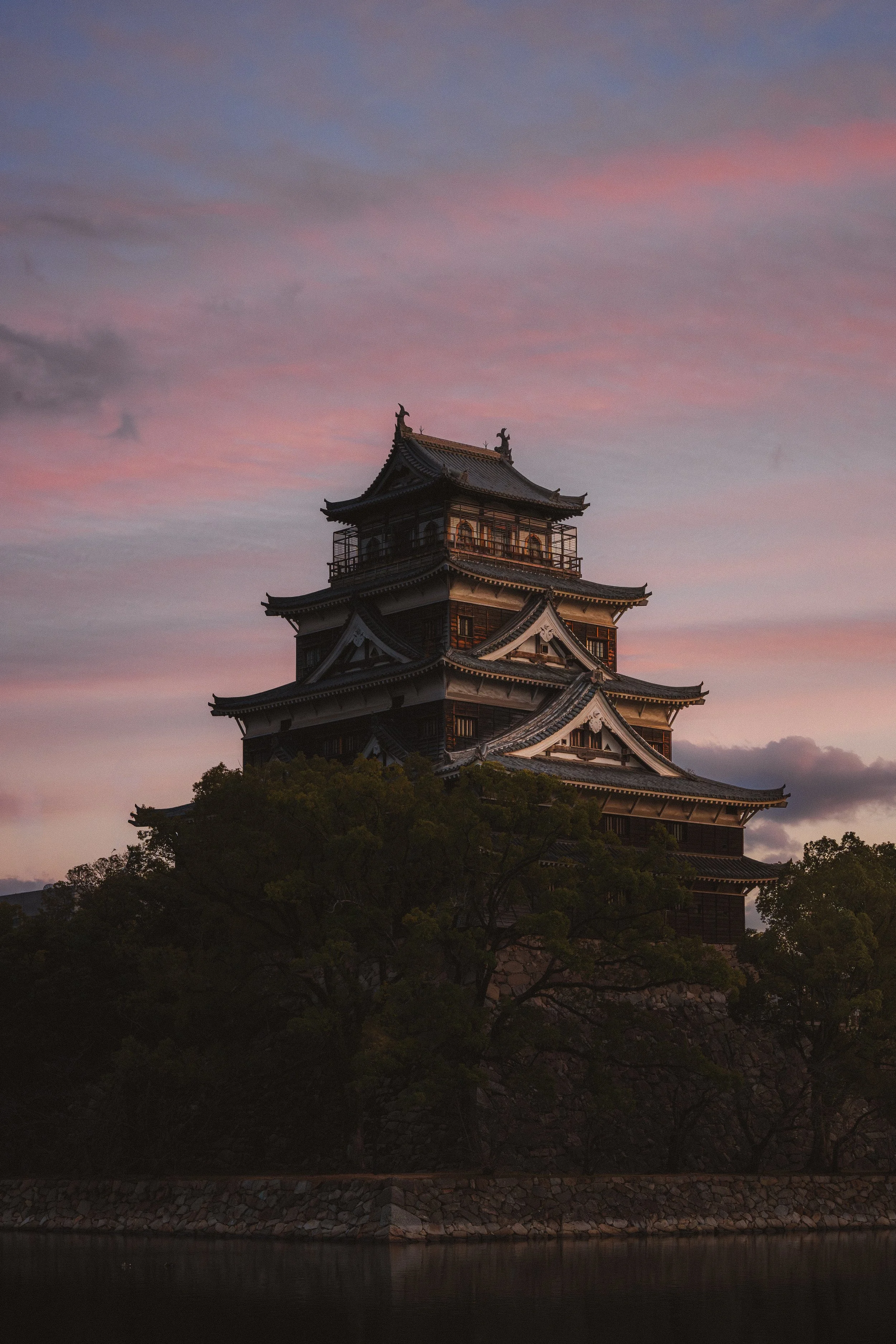Japanese castle at sunset with pink and purple sky, surrounded by trees and positioned near a body of water.