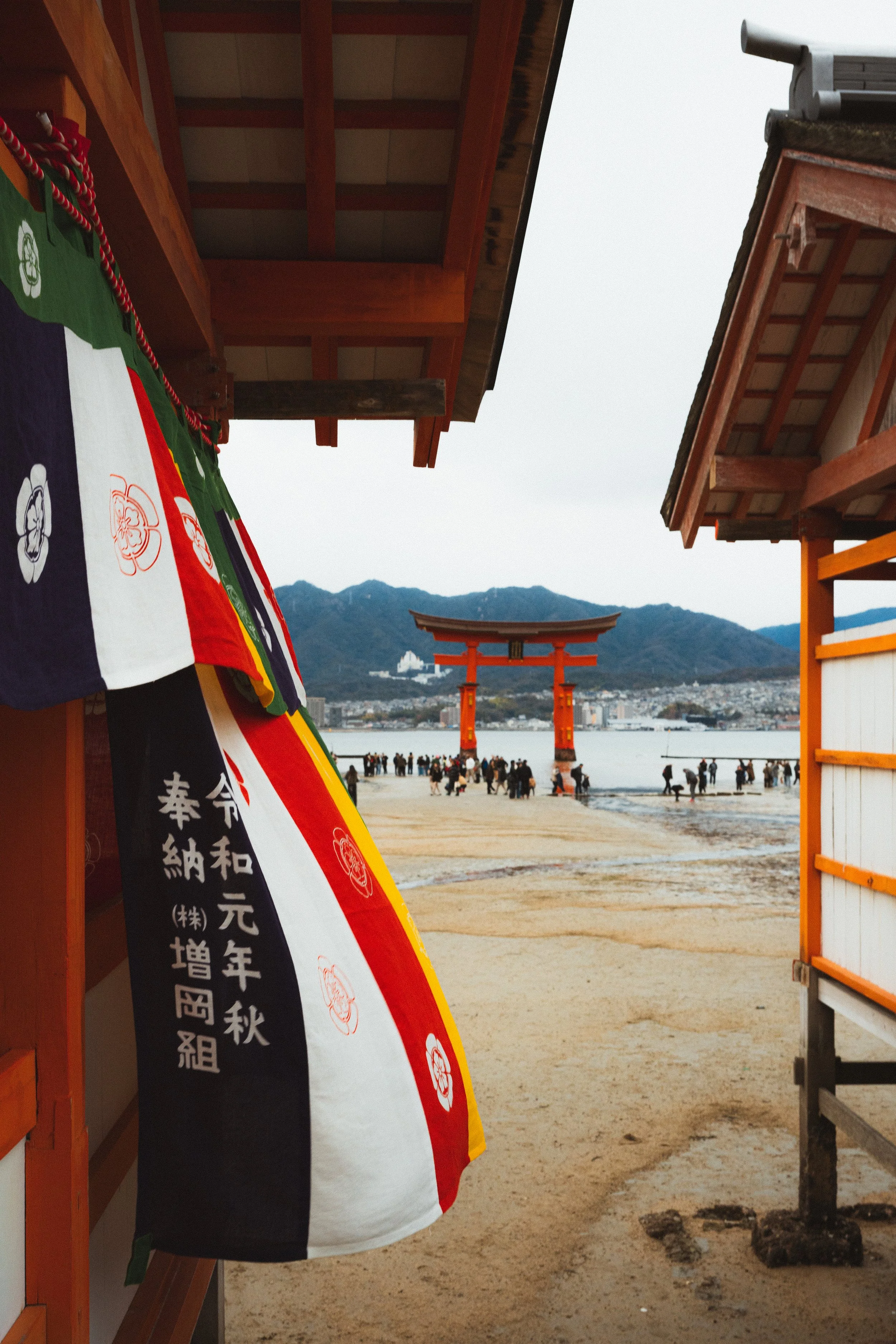 View of a traditional Japanese torii gate at a beach, with mountains in the background and a cloudy sky. The photo is taken from a shaded area with buildings and a colorful banner on the side.