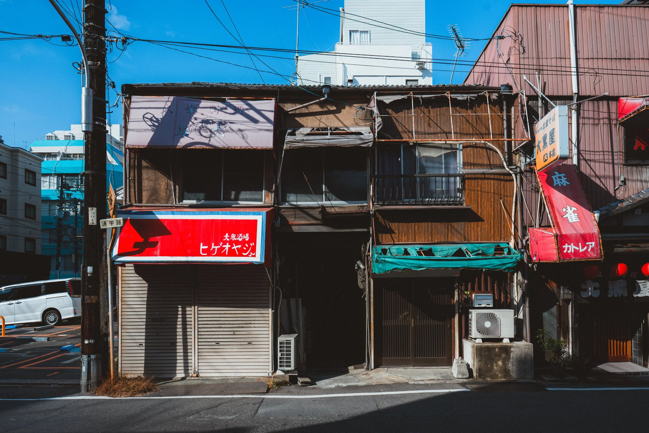 Shot of an urban street scene with a closed storefront, a parking lot with cars, and a utility pole with wires. Buildings and clear blue sky in the background.