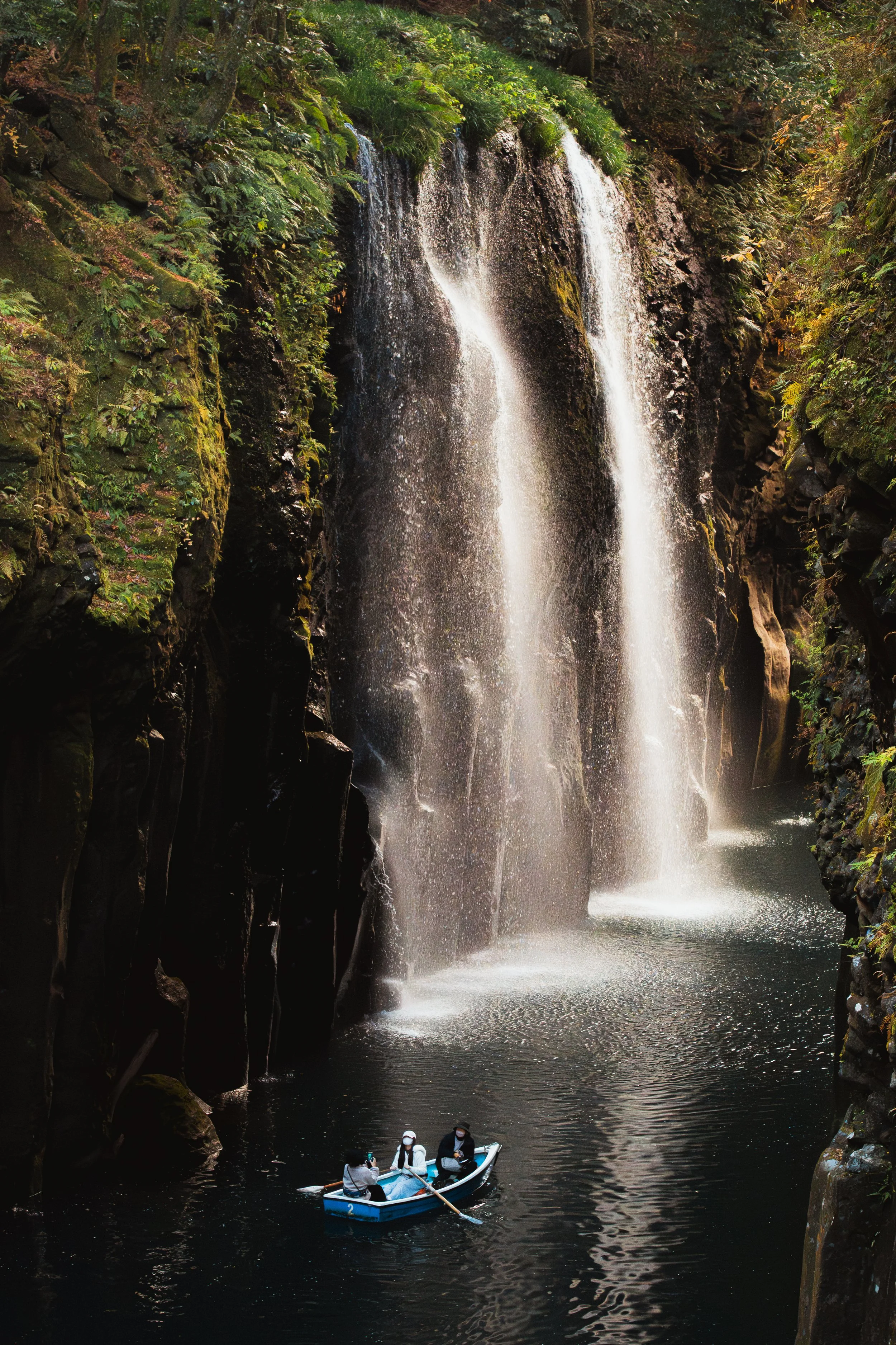 People in a boat rowing beneath a waterfall in a lush, green canyon.