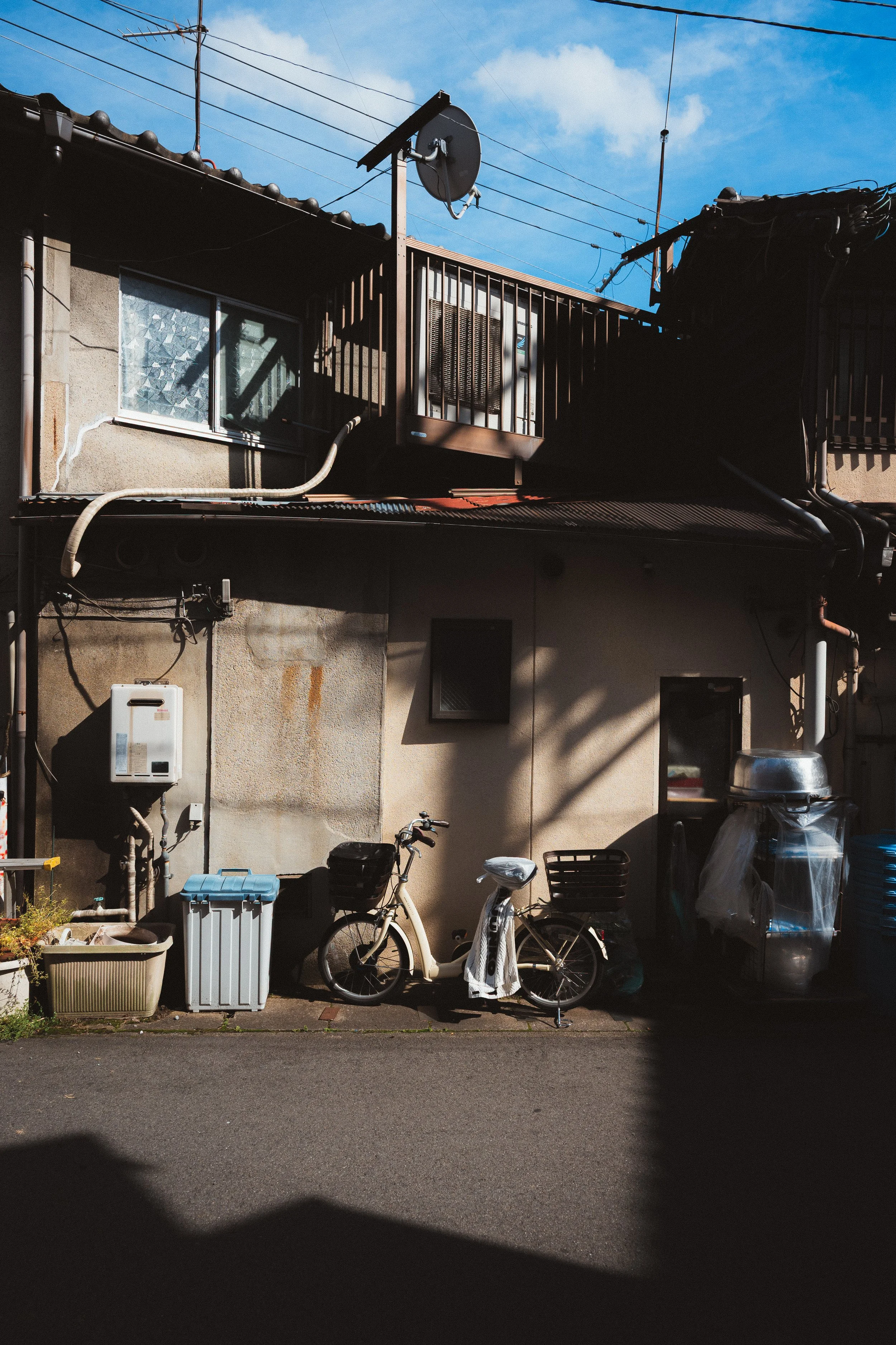 Backyard scene with bicycles, trash bins, plants, and a building with windows and an air conditioning unit, under a blue sky with some clouds.