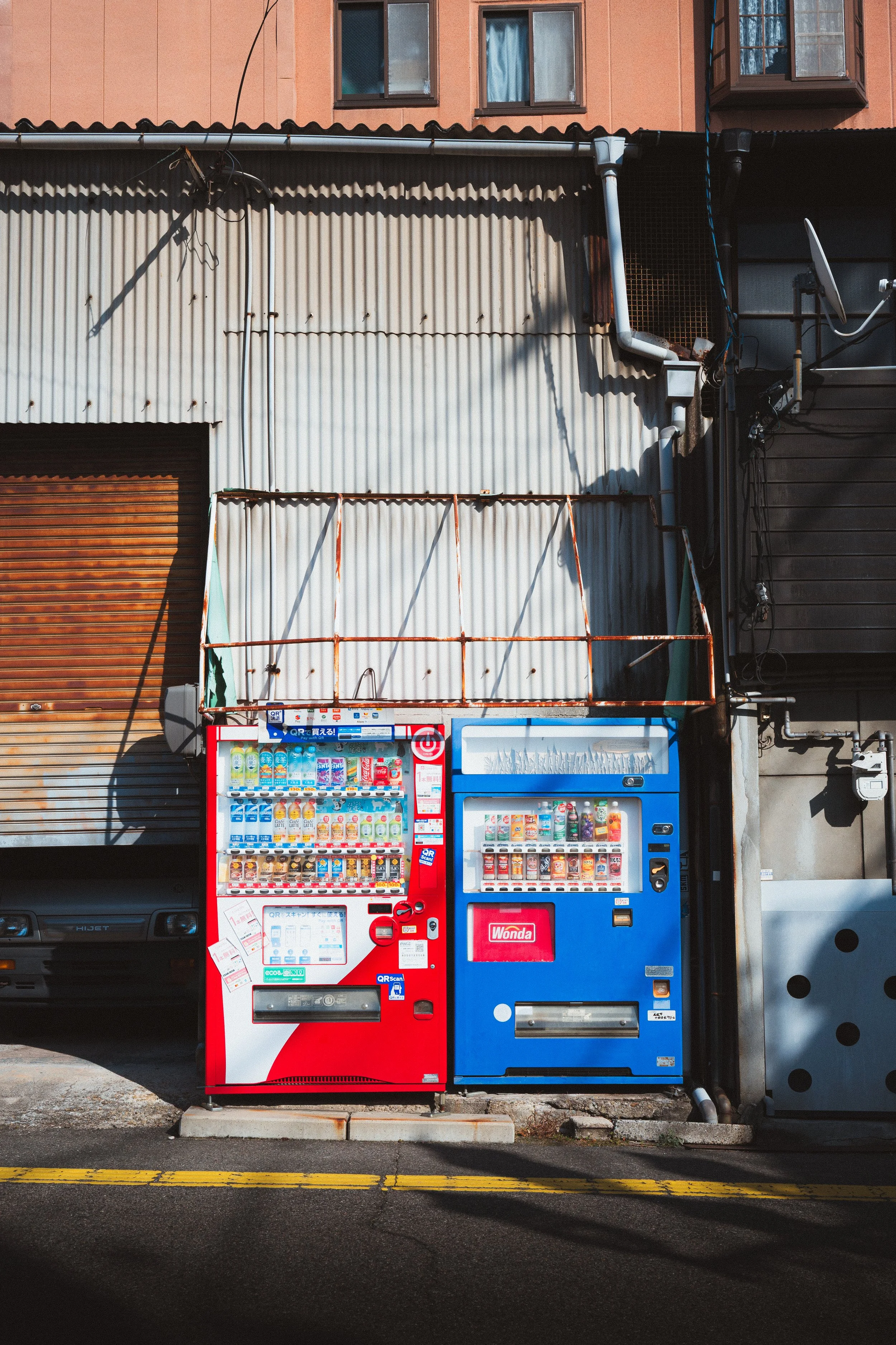 Two vending machines, one red and one blue, stand side by side on a sidewalk in front of a building with an orange upper level and beige corrugated metal lower siding. Shadows and wires are visible on the building's facade.