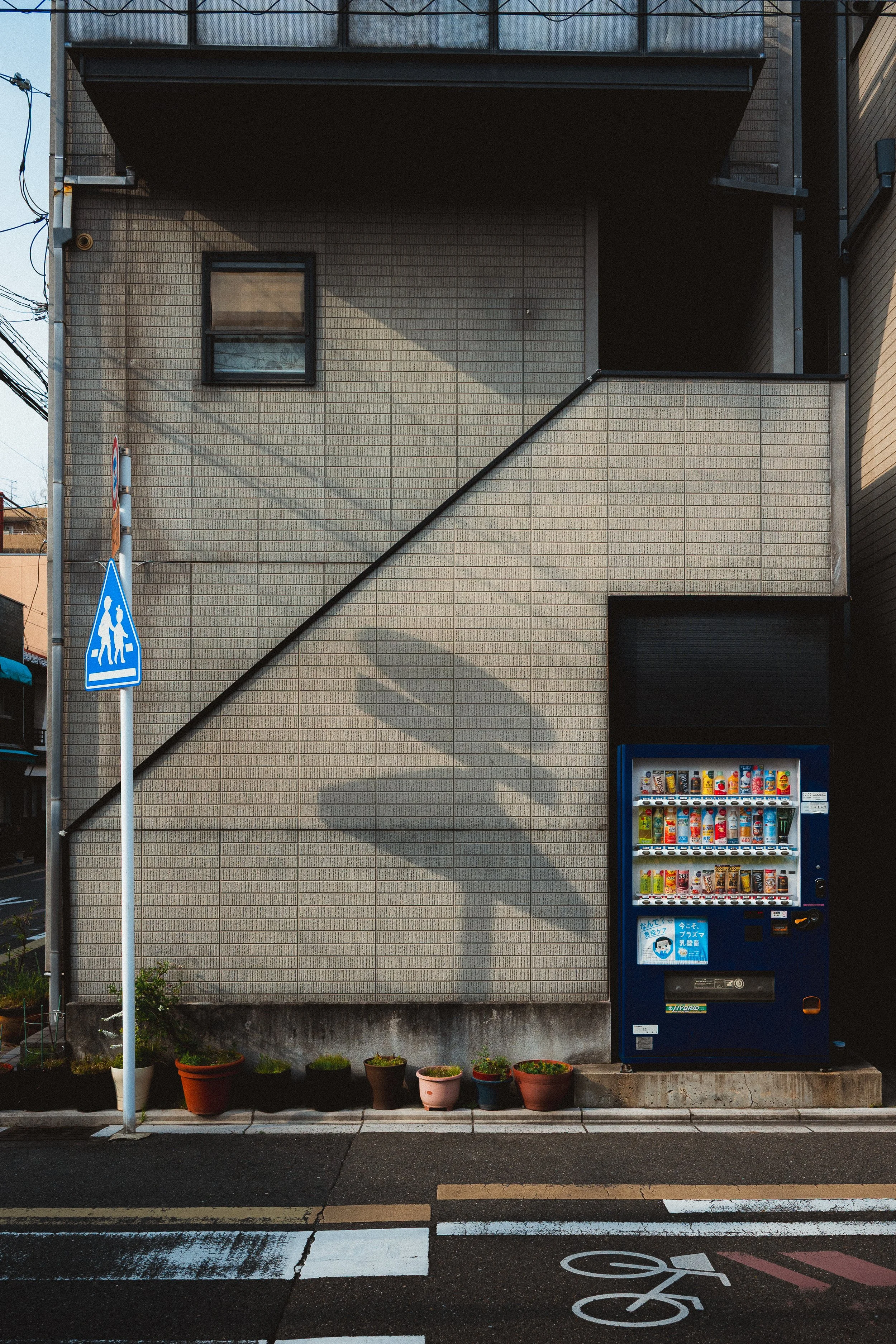 A building with beige brick walls, small window, potted plants at the base, a vending machine on the right, a pedestrian crossing sign, and a bicycle lane mark on the street.