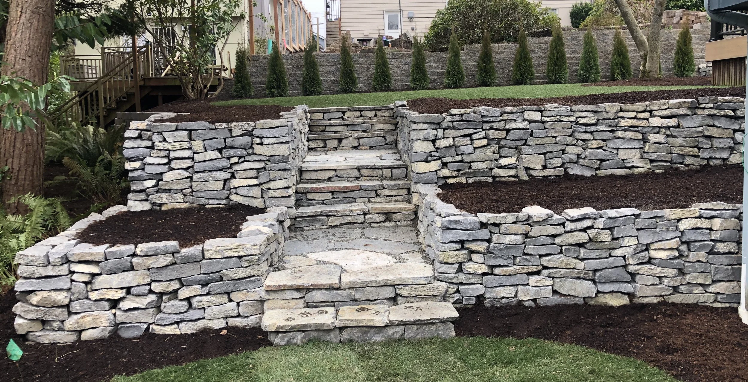 Tiered backyard garden with stone retaining walls, wooden stairs, and landscaped areas with plants and grass.
