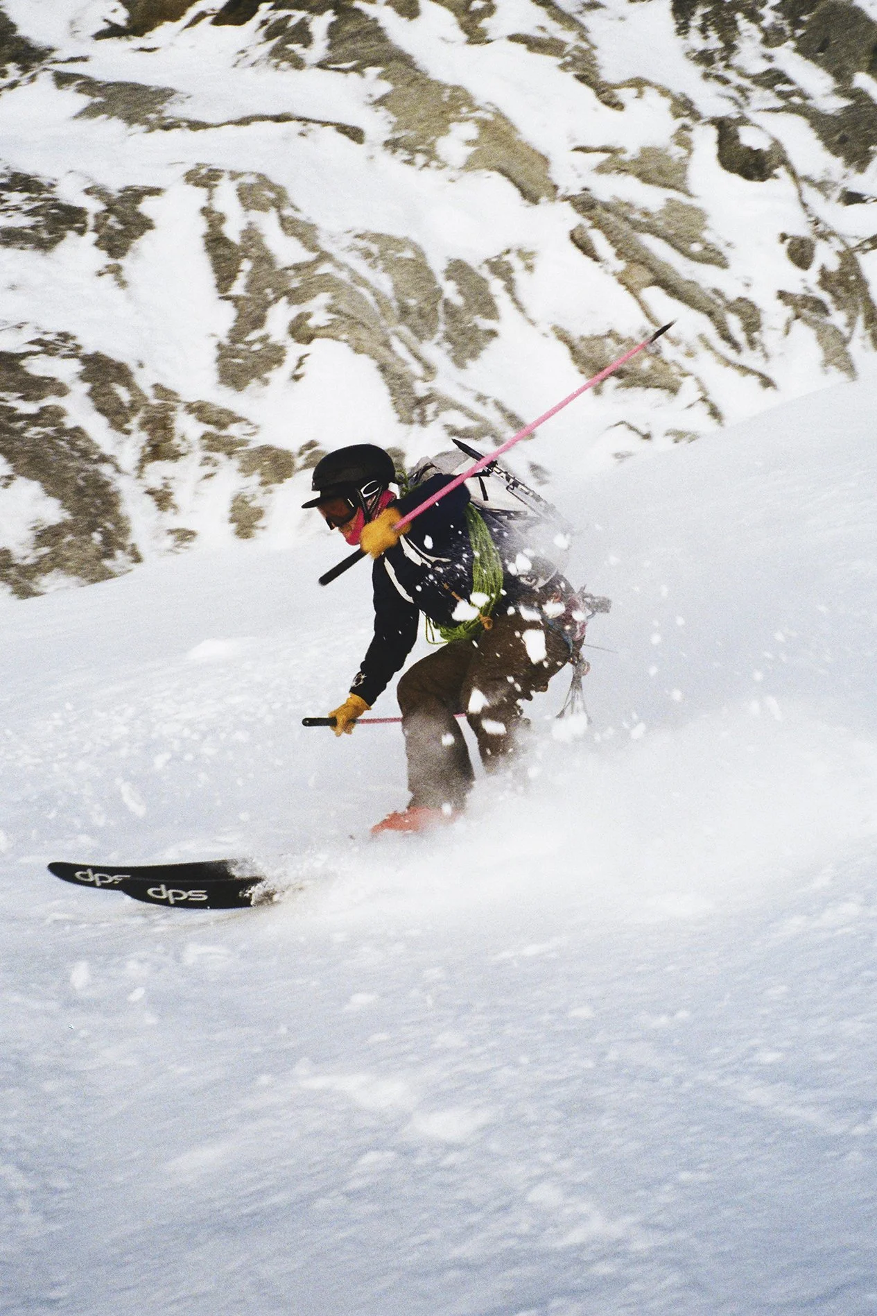 A person wearing a helmet, goggles, a backpack, and gloves is skiing down a snowy slope in a mountainous area with rocky peaks in the background.