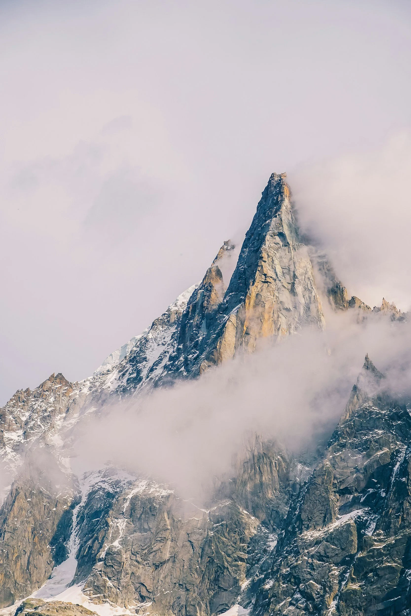 Snow-covered mountain peak with steep rocky slopes, surrounded by mist and clouds.