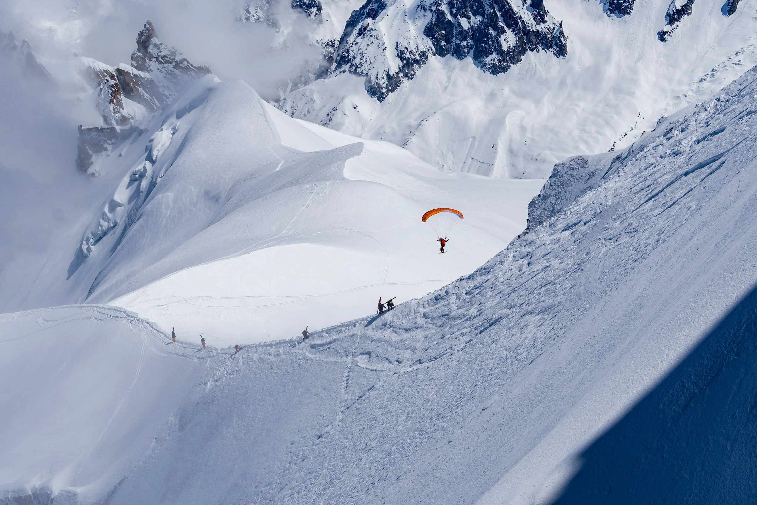 A snowy mountain scene with a person paragliding above snow-covered slopes, other skiers and snowboarders on the ground, and steep mountain peaks in the background.