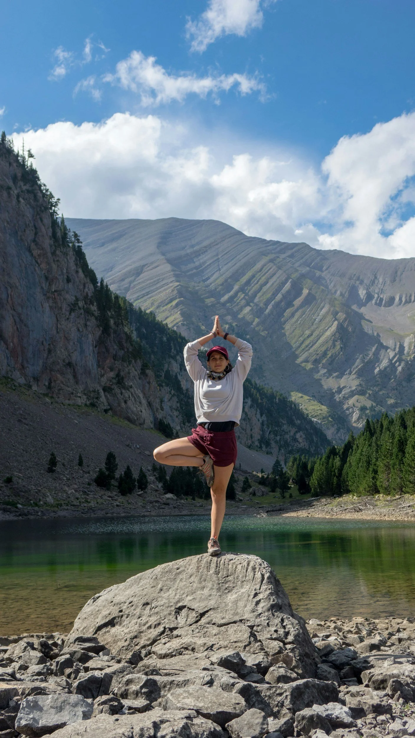 A woman practicing yoga on a large rock beside a lake with mountains and trees in the background.
