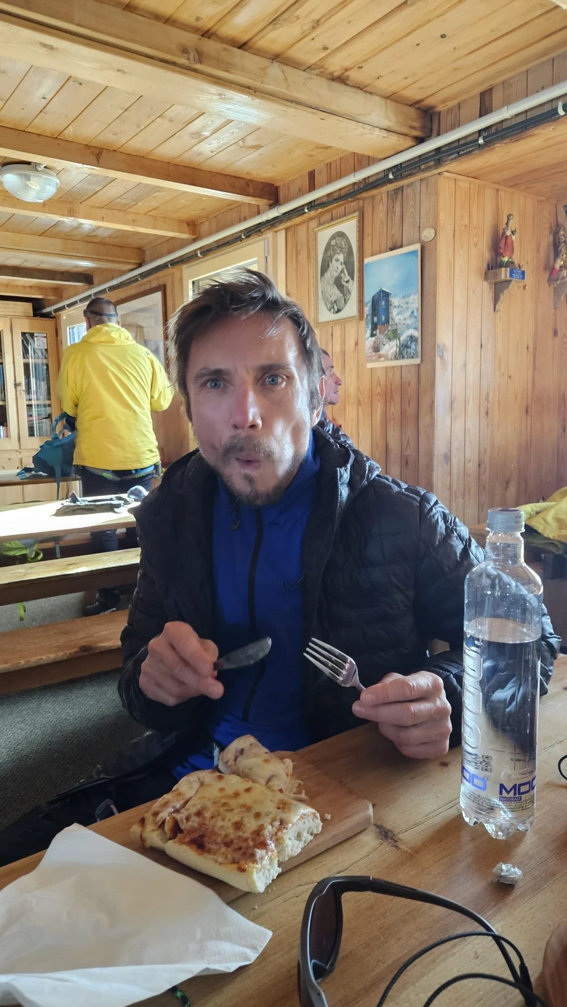 A man with blue eyes and a beard, wearing a blue jacket and black vest, is sitting at a wooden table inside a cabin-style restaurant. He is holding a fork in his right hand and a knife in his left hand, with a slice of pizza on the table in front of him. There is a plastic water bottle on the table. In the background, there are wooden walls with framed pictures and a man in a yellow jacket standing.