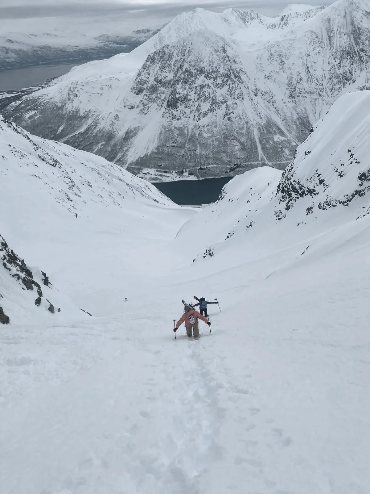 Two mountaineers trekking through snow in a mountainous landscape with icy peaks and a lake in the distance.