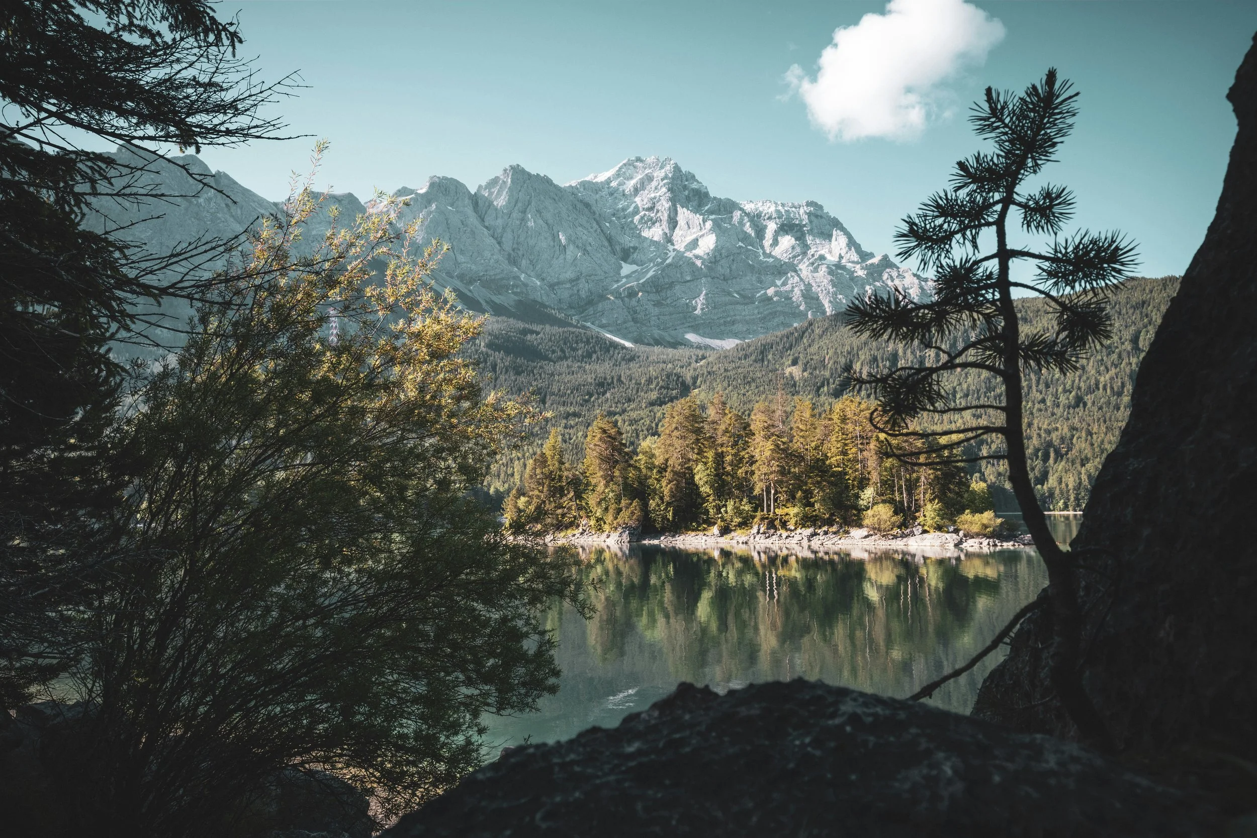 A scenic mountain landscape with tall peaks, a forest, and a calm lake in the foreground, with trees framing the view and a blue sky with a few clouds.