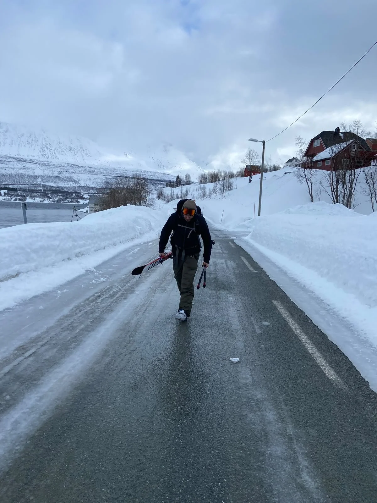 Person walking on a snow-covered road carrying skis and walking poles in a snowy landscape with houses and mountains in the background.