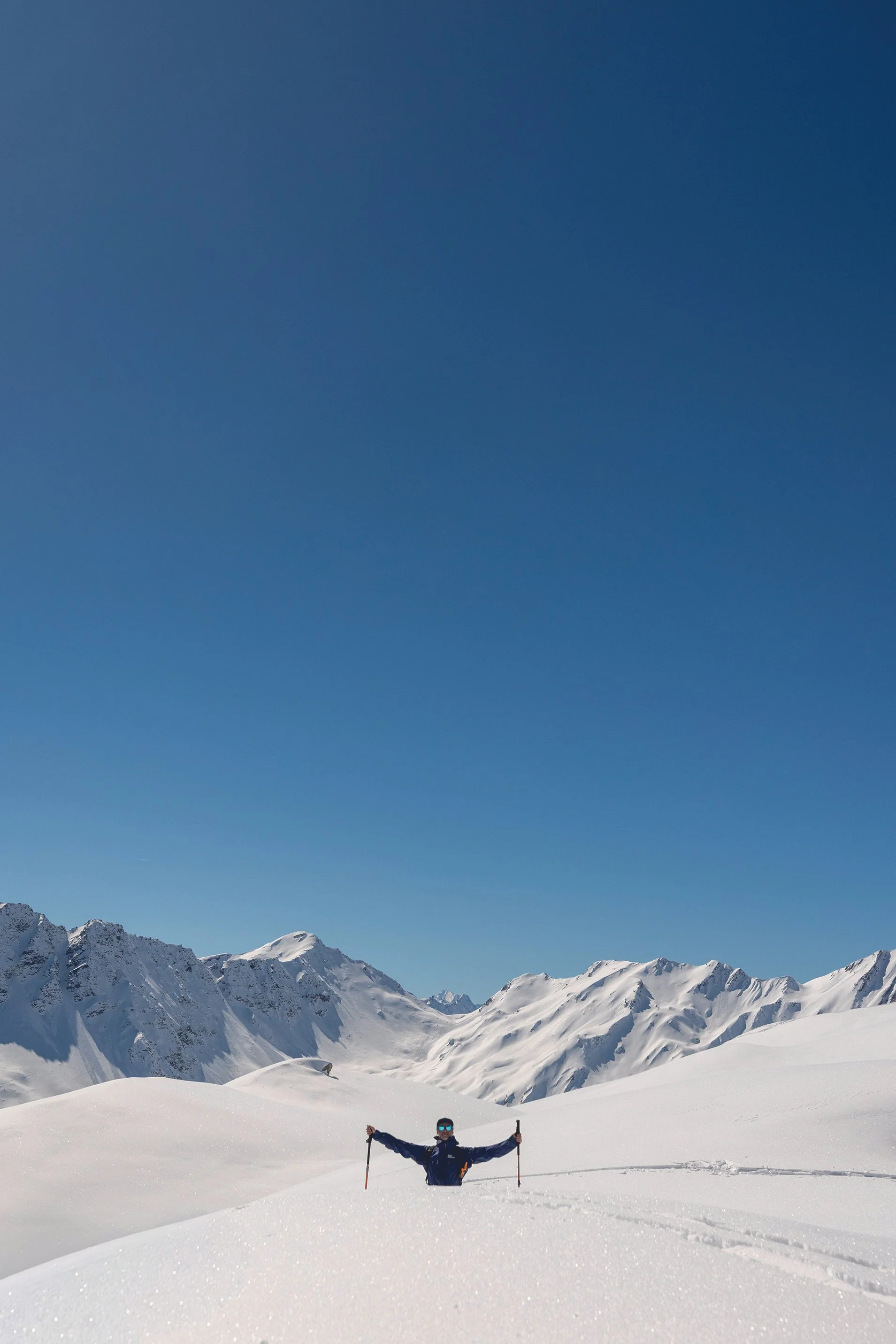 Person standing in snowy mountain landscape with arms outstretched, holding ski poles, and surrounded by snow-covered peaks under clear blue sky.