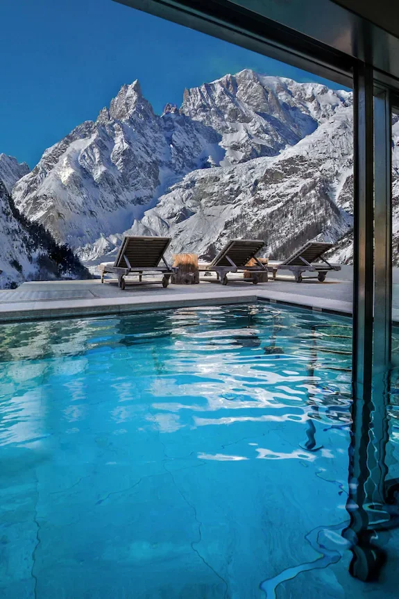Indoor pool with large glass window showing snow-covered mountains and blue sky outside, with three lounge chairs on the deck.