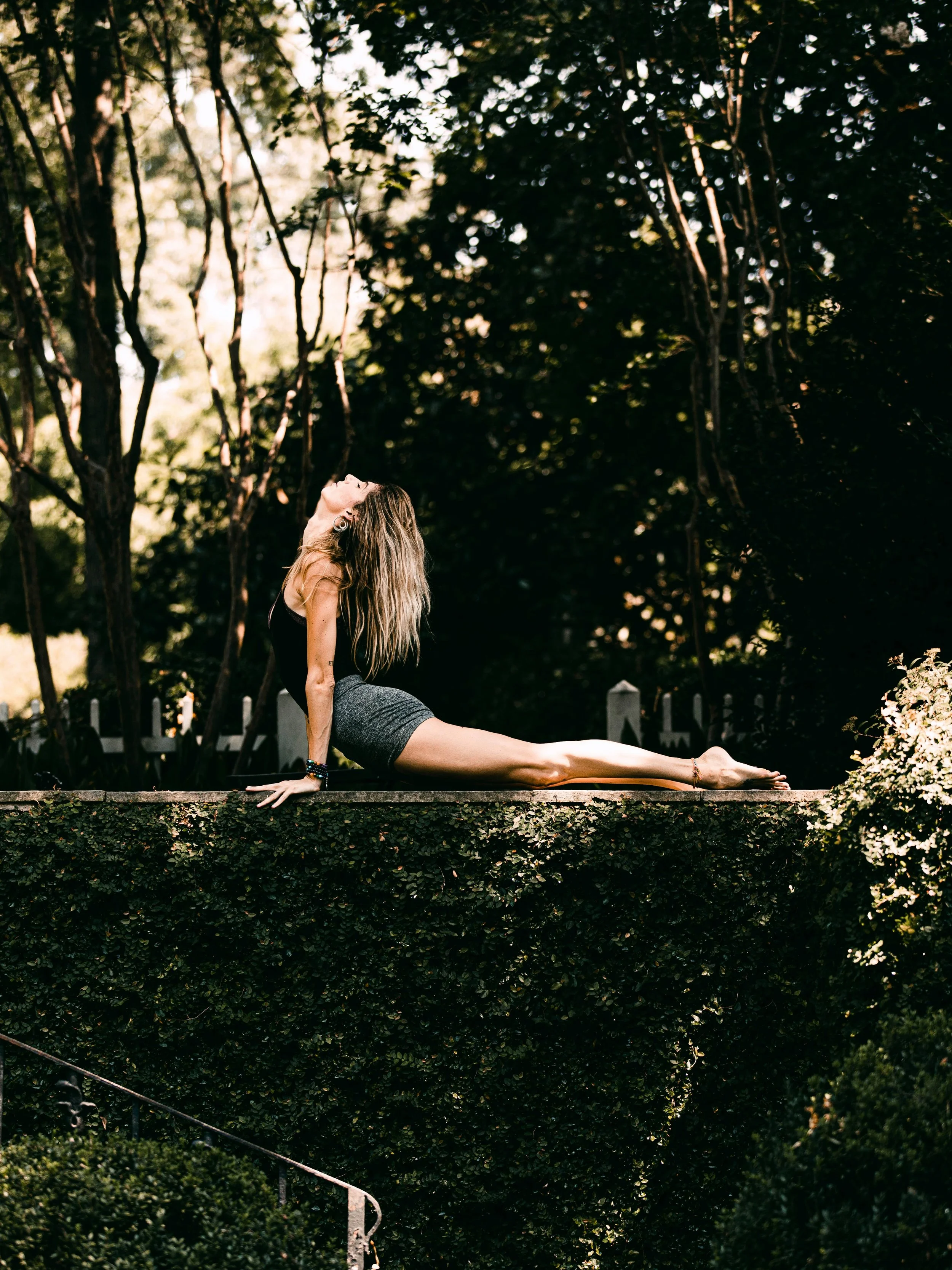 A woman practicing yoga outdoors on a stone ledge in a lush, green park, performing the Cobra pose with her head tilted back and arms extended behind her.