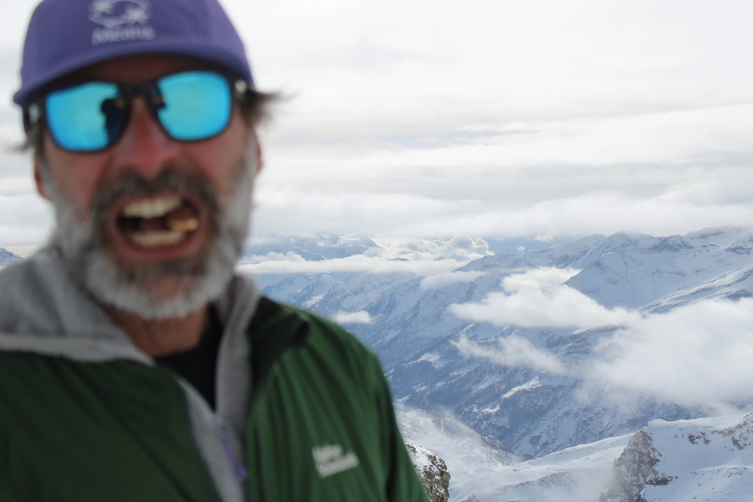 A man with a gray beard, wearing sunglasses, a purple cap, and a green jacket, smiles in front of a snowy mountain landscape with clouds and peaks in the background.