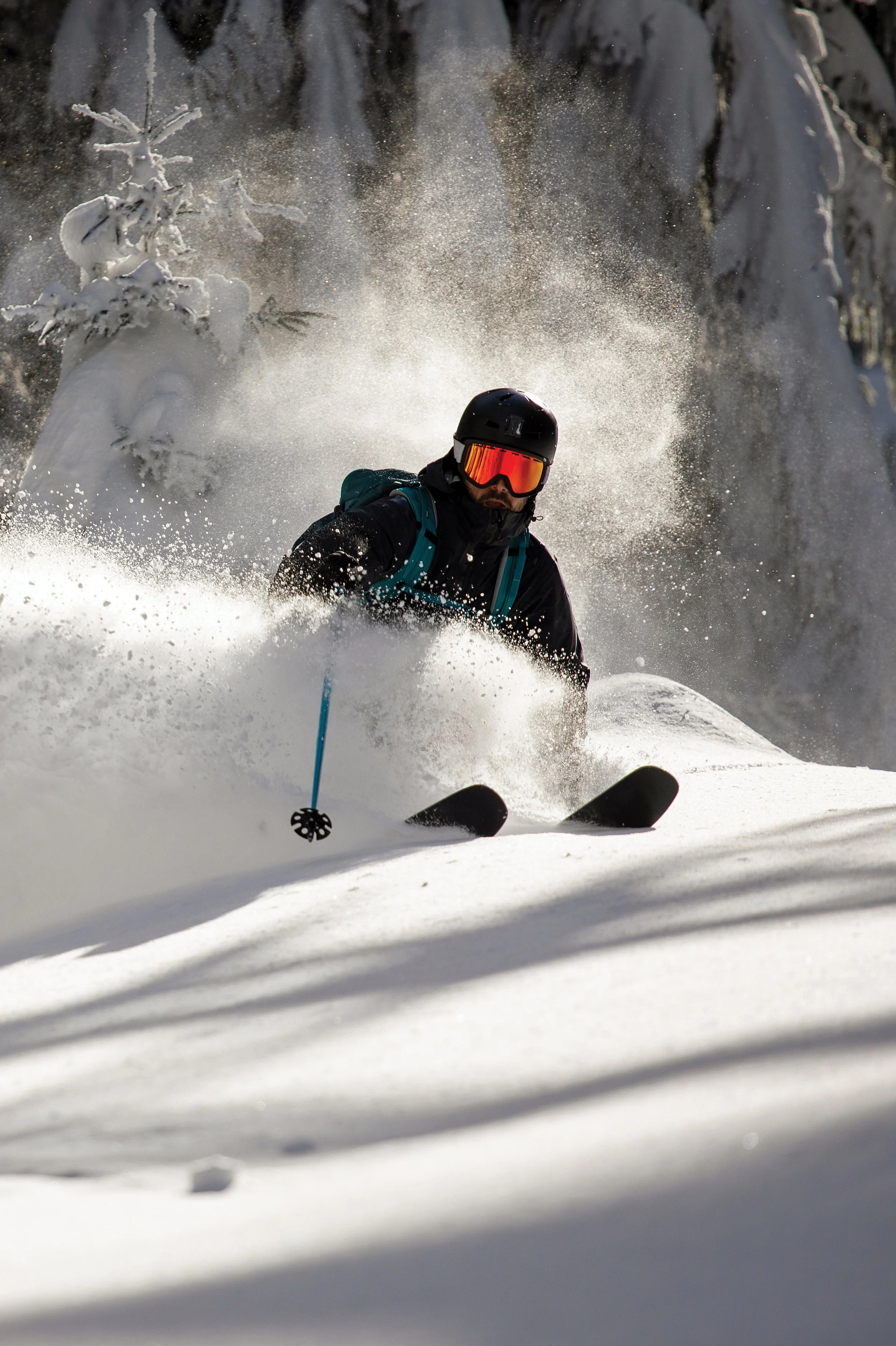 A skier in black gear and a helmet descending a snowy mountain, surrounded by snow-covered trees, with snow spraying up around him.