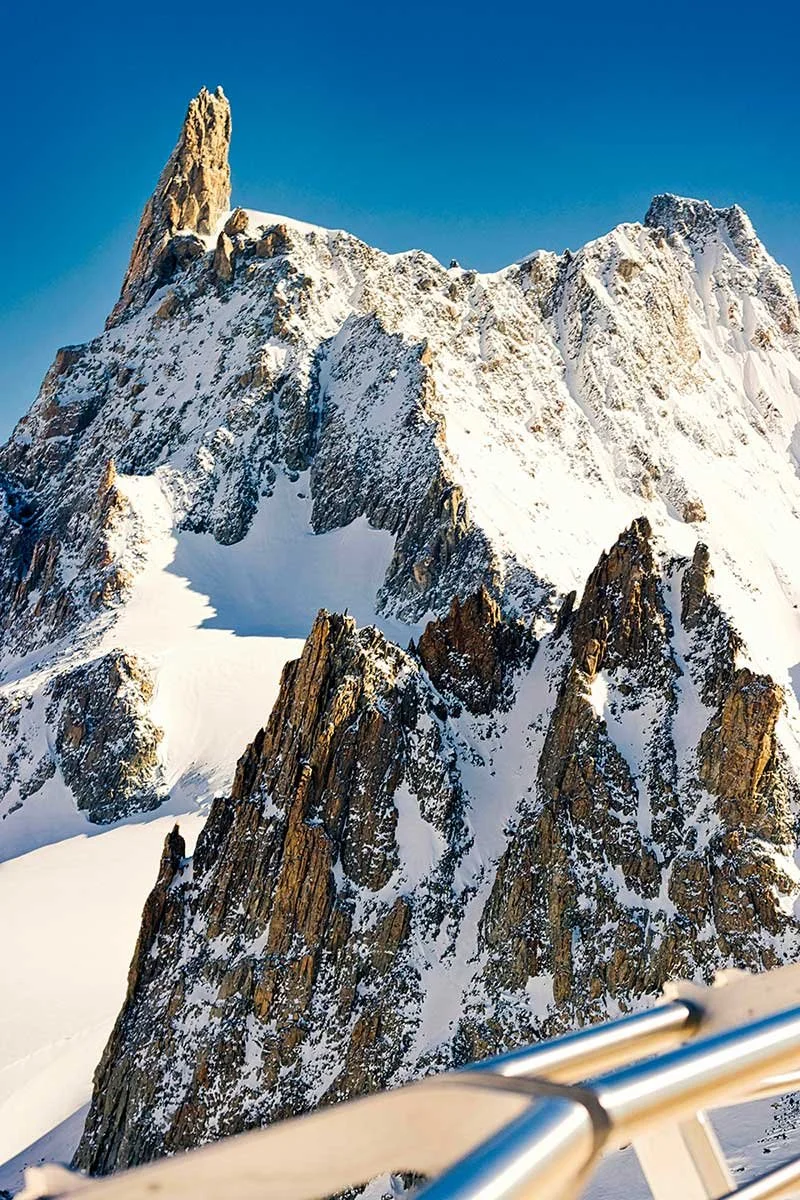 Snow-covered jagged mountain peaks against a clear blue sky, seen from a viewing platform with a metal railing.