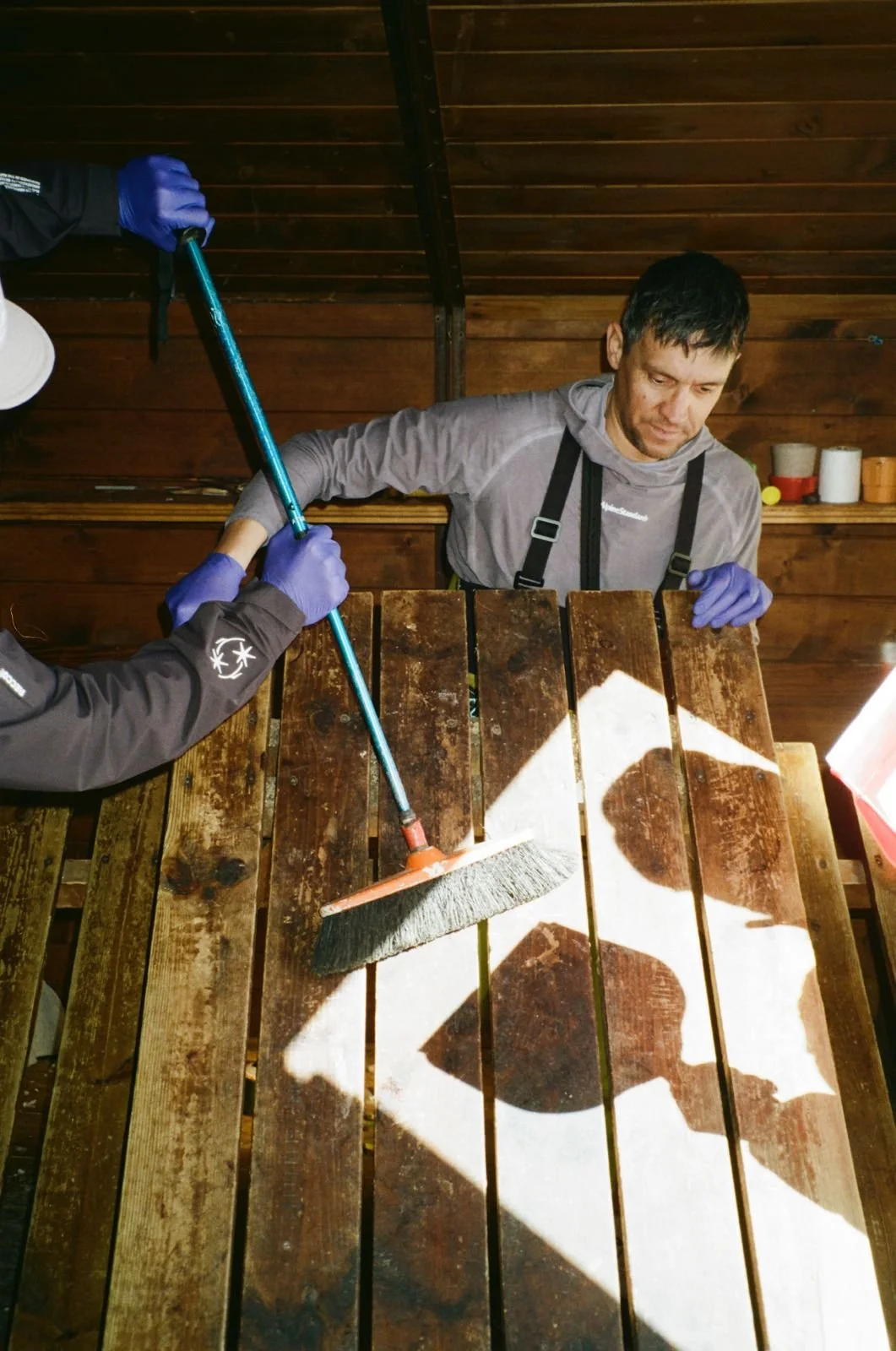 Person cleaning a wooden table with a broom while another person holds the handles, both wearing gloves, in a wooden-paneled room.
