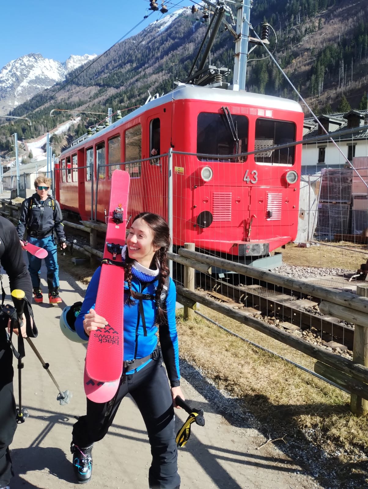 A woman holding a pink snowboard with a group of people in front of a red train car at a mountain station, with snow-covered mountains in the background.