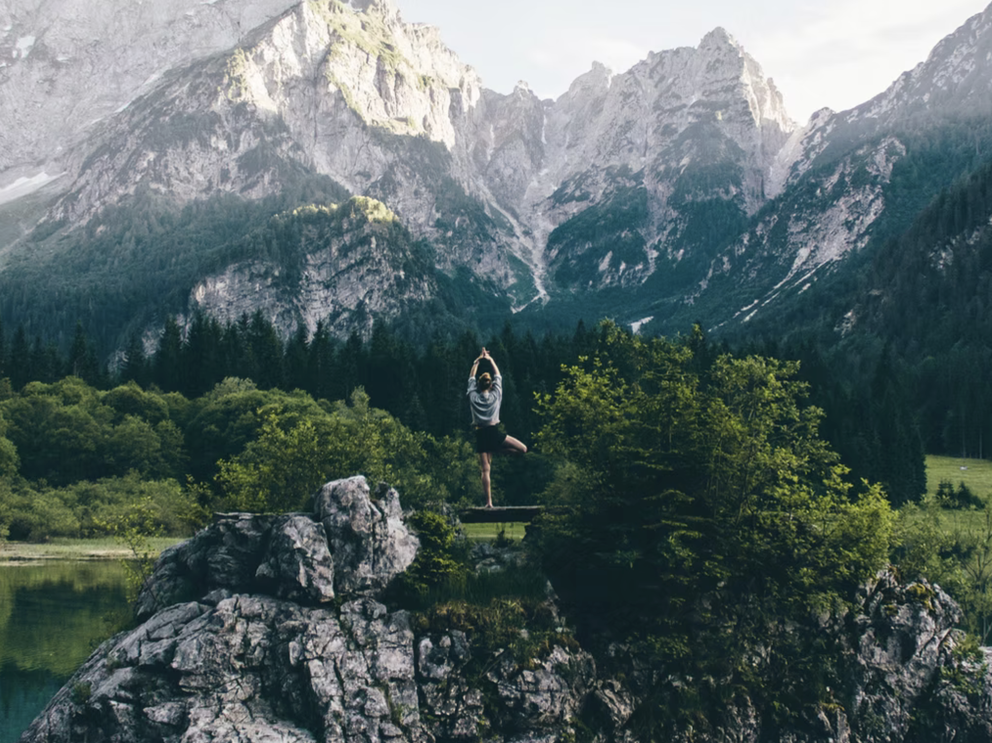 Person practicing yoga on a rock by a lake with a backdrop of towering mountain peaks and forested hills.