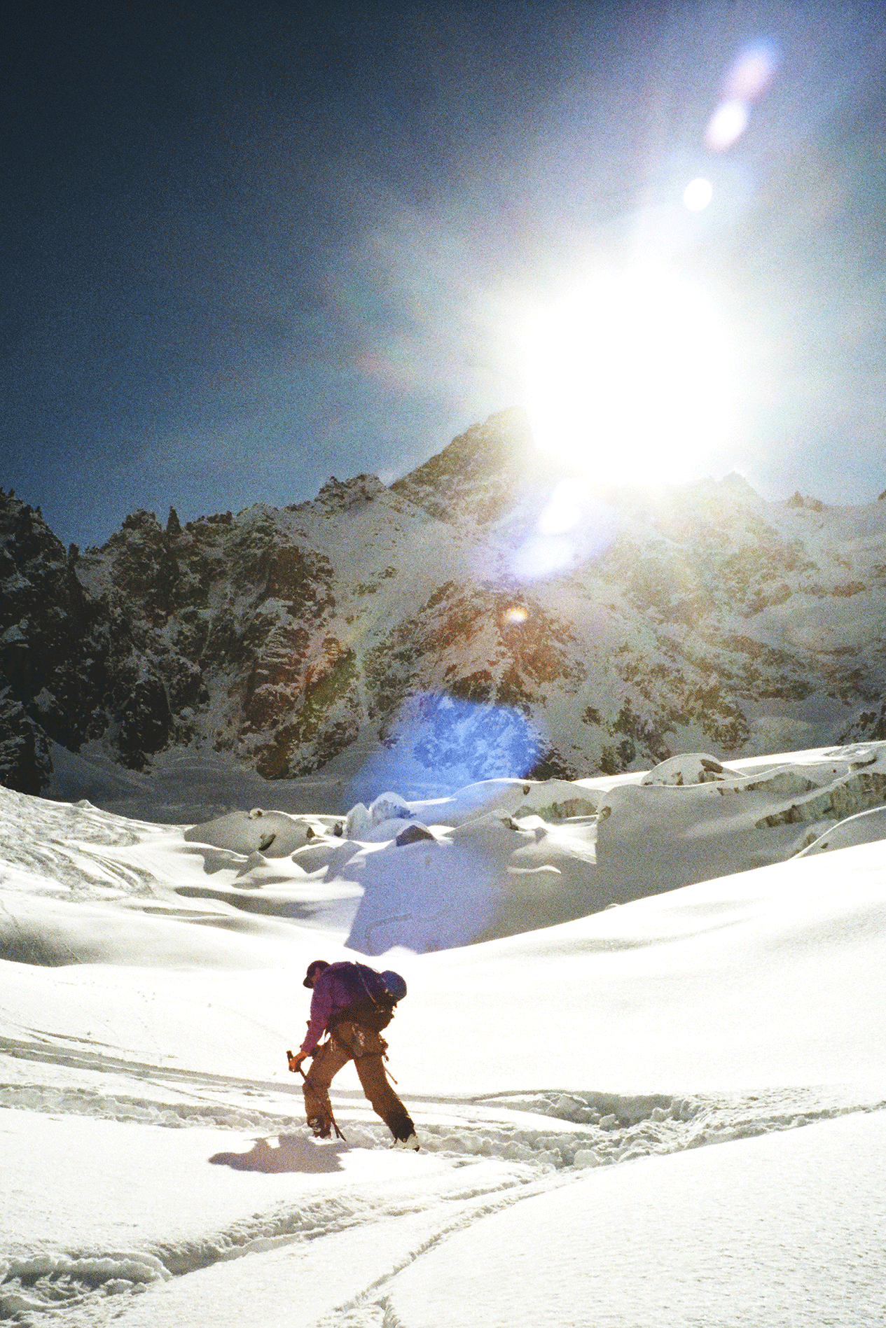 A person skiing or snowboarding in a snowy mountain landscape under a bright sun with a clear sky.