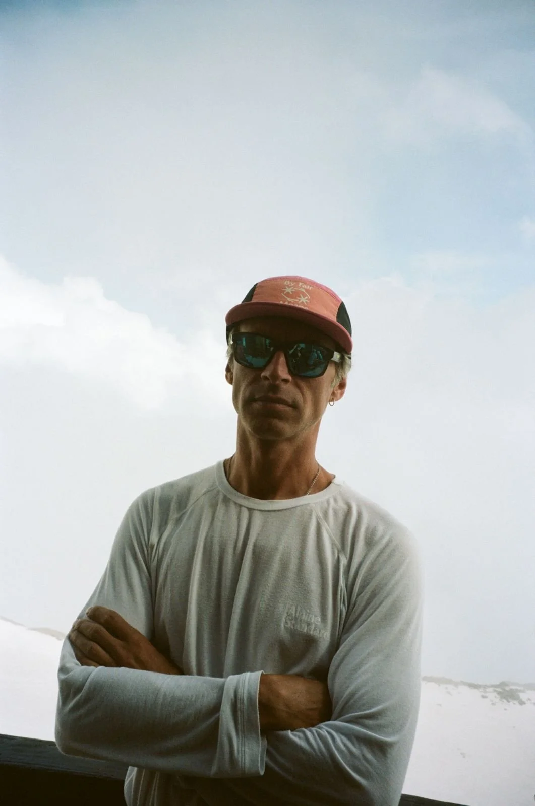 A man standing outdoors against a cloudy sky, wearing a baseball cap, sunglasses, a white long-sleeve shirt, and an earring.