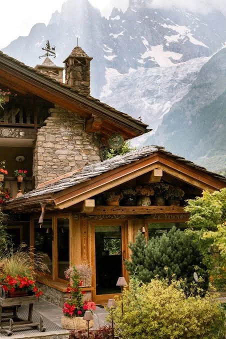 A rustic mountain cabin with stone and wood architecture, surrounded by greenery, with snow-capped mountains in the background.