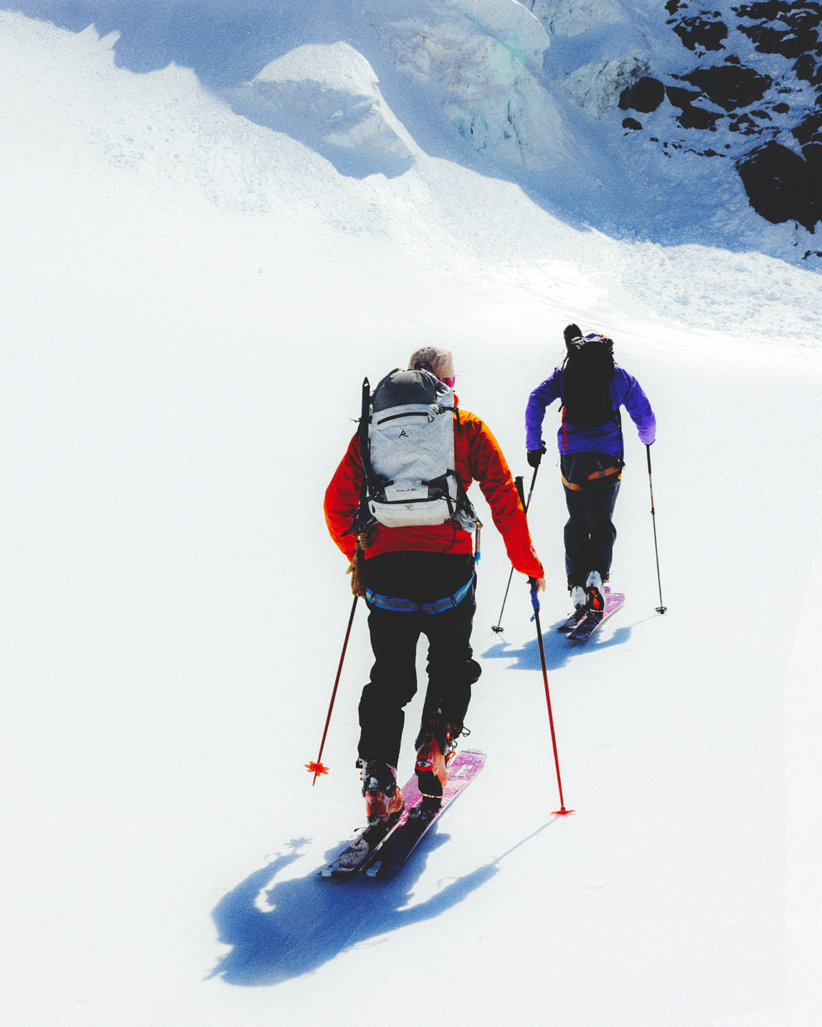 Two people in winter gear and backpacks skiing uphill on a snow-covered mountain landscape.