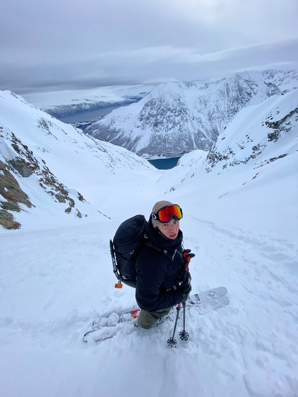 A man in ski gear, sunglasses, and a beanie kneeling on snowy mountain slopes with a backpack, surrounded by snow-covered mountains and a body of water in the distance.