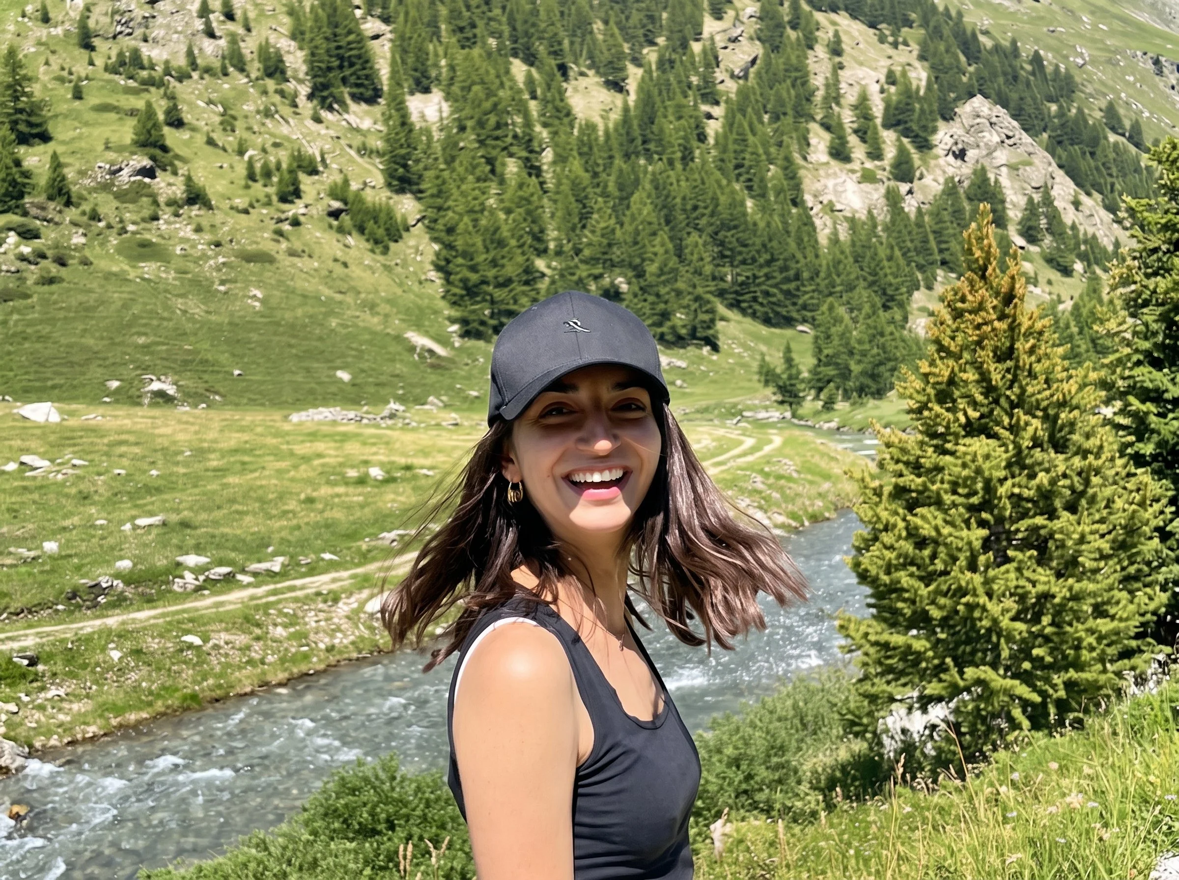 A woman smiling outdoors in a mountainous area with green trees, rocky hills, and a river behind her, wearing a black cap and sleeveless top.