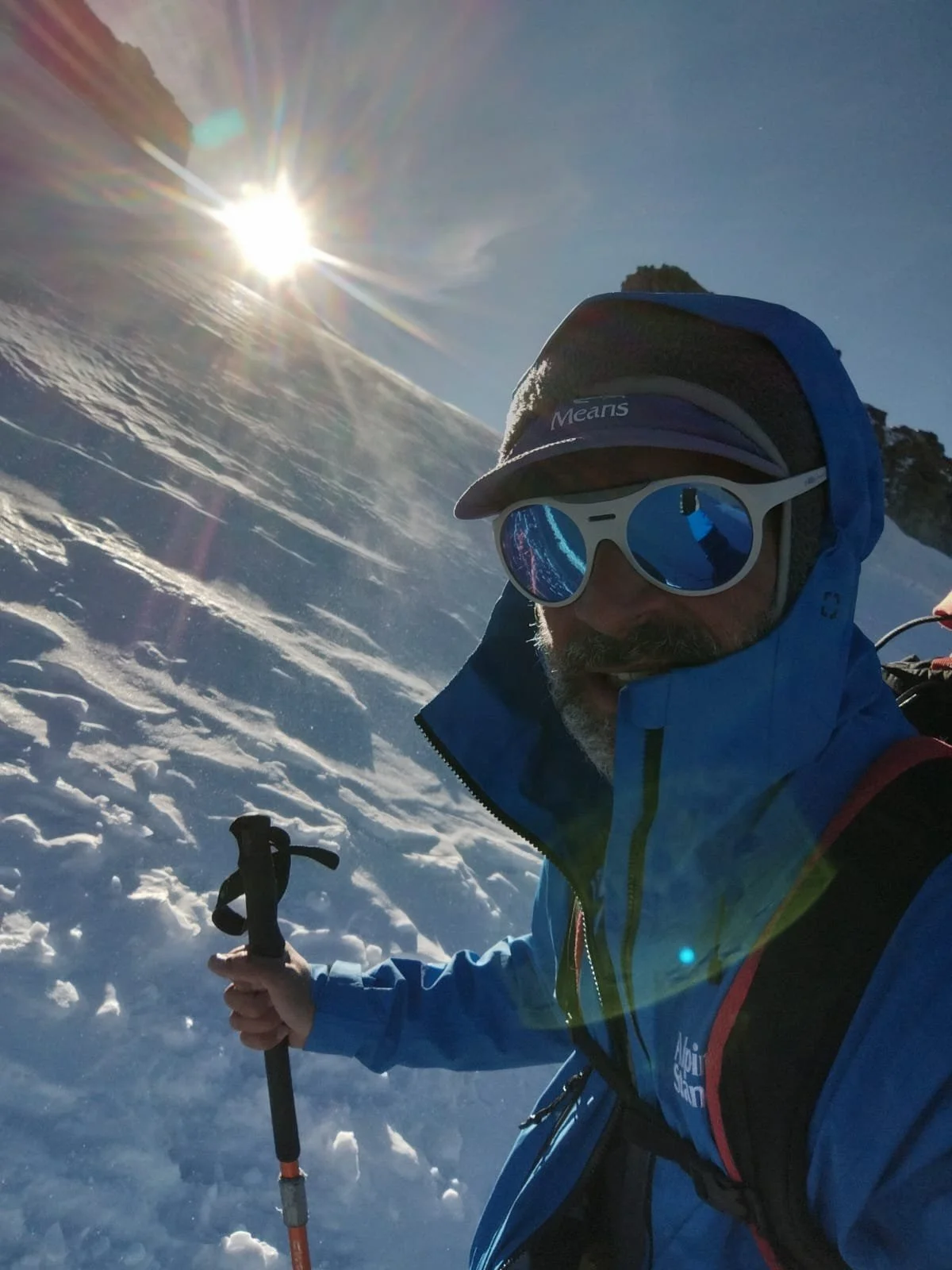 A man dressed in winter gear, including a blue jacket, sunglasses, and a cap, takes a selfie in a snowy mountain landscape with the sun shining brightly in the sky.