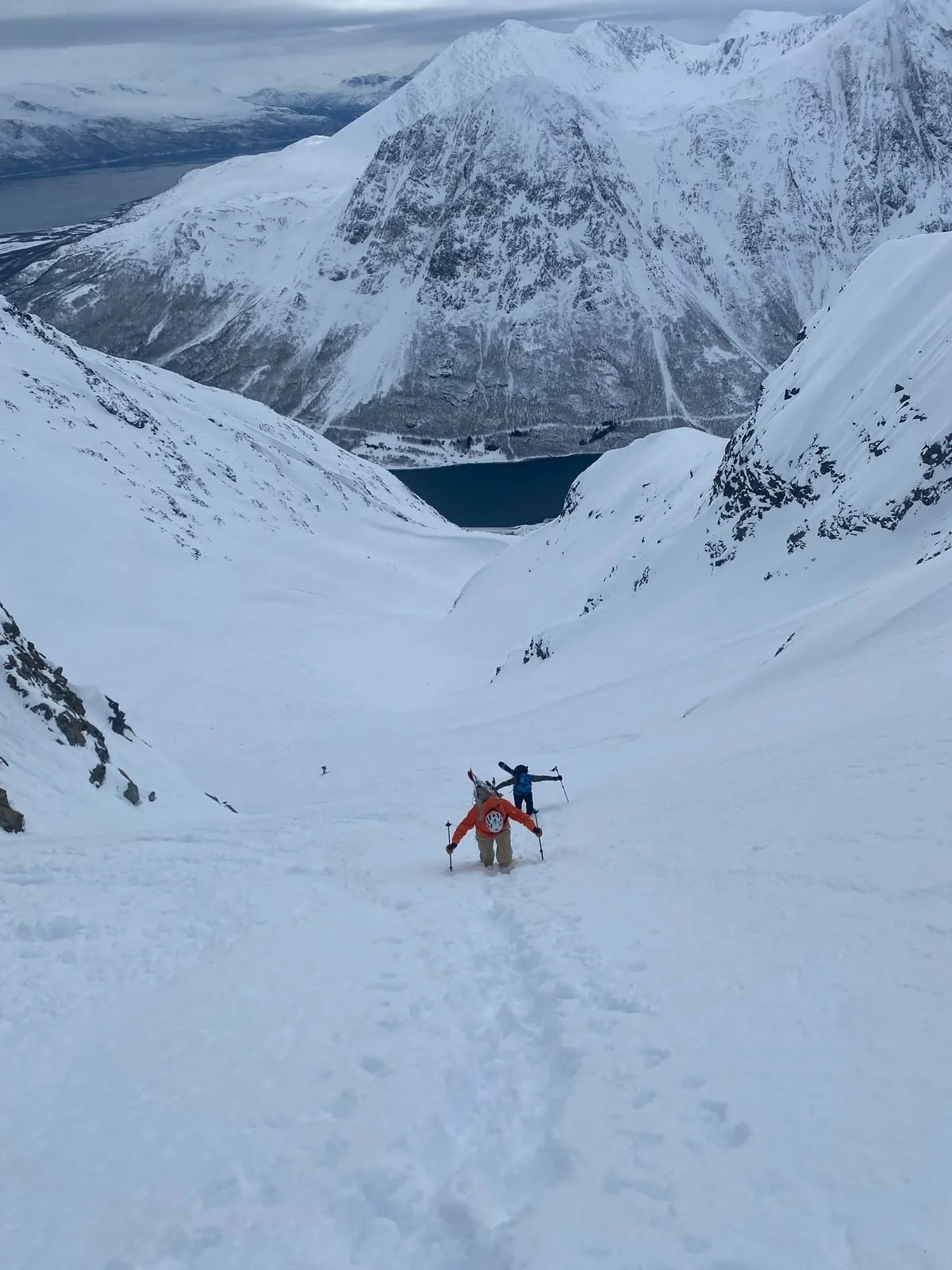 Two hikers climb a snowy mountain slope with a scenic backdrop of snow-covered peaks and a fjord below, during daytime.