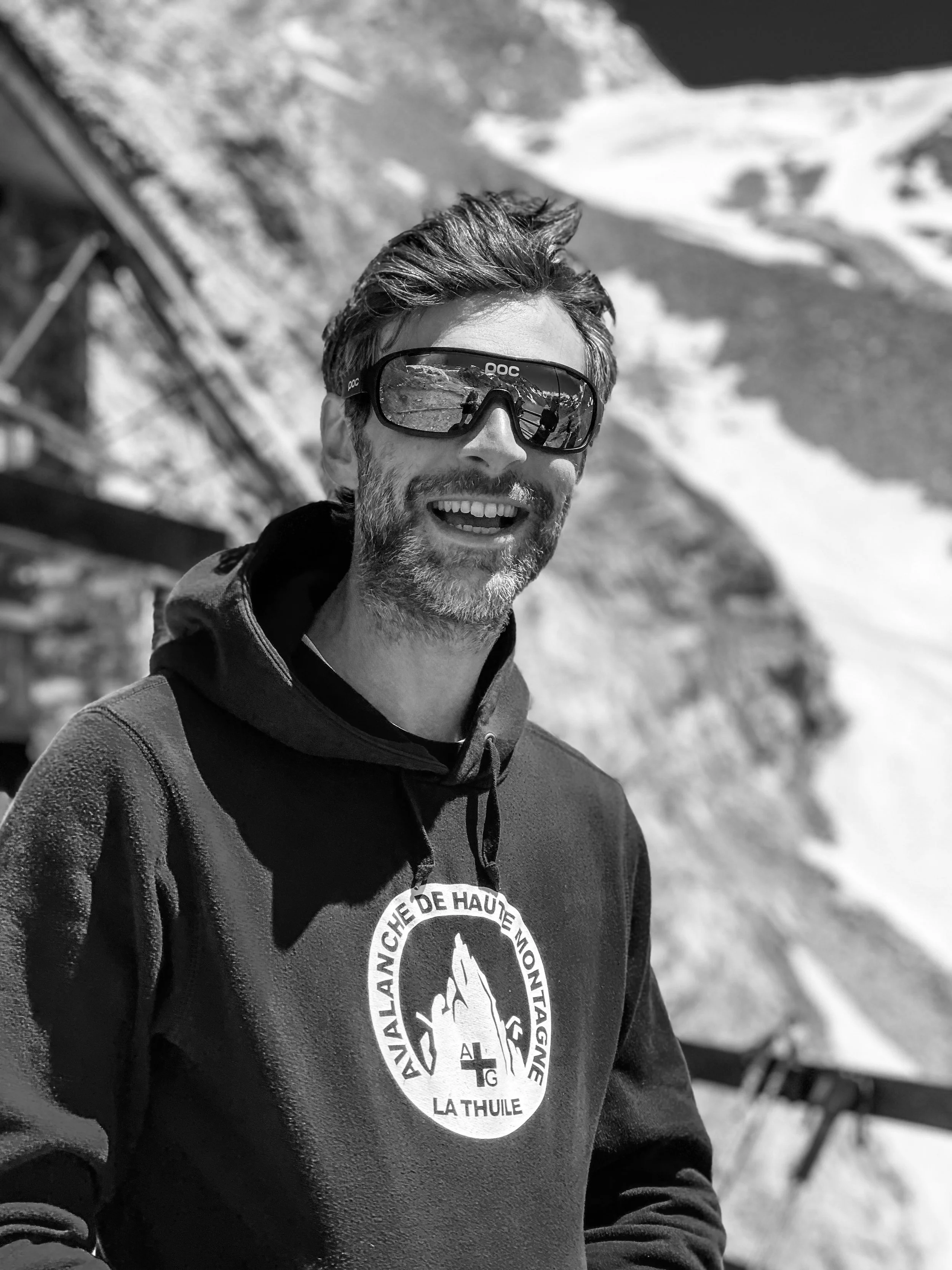 A smiling man with sunglasses and a beard, wearing a hoodie with a logo, taking a selfie outdoors in a snowy mountainous area.