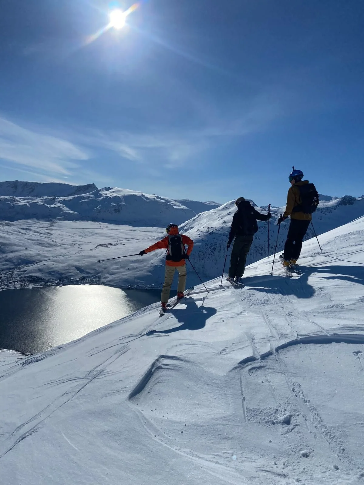 Three people skiing on a snowy mountain ridge with a snow-covered landscape and mountain range in the background under a clear blue sky and bright sun.