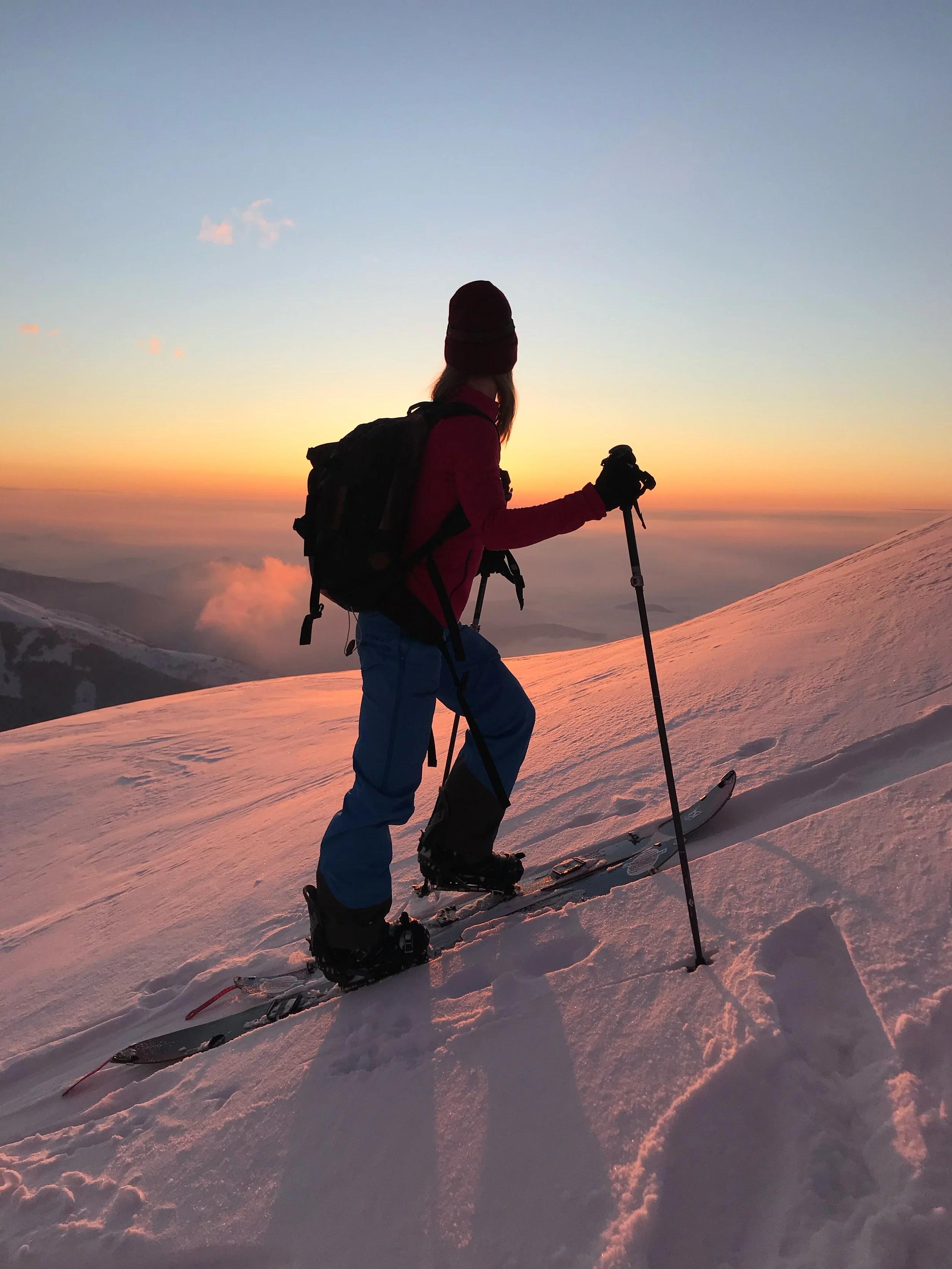 Person in winter gear with backpack and ski poles standing on a snowy mountain slope at sunset.