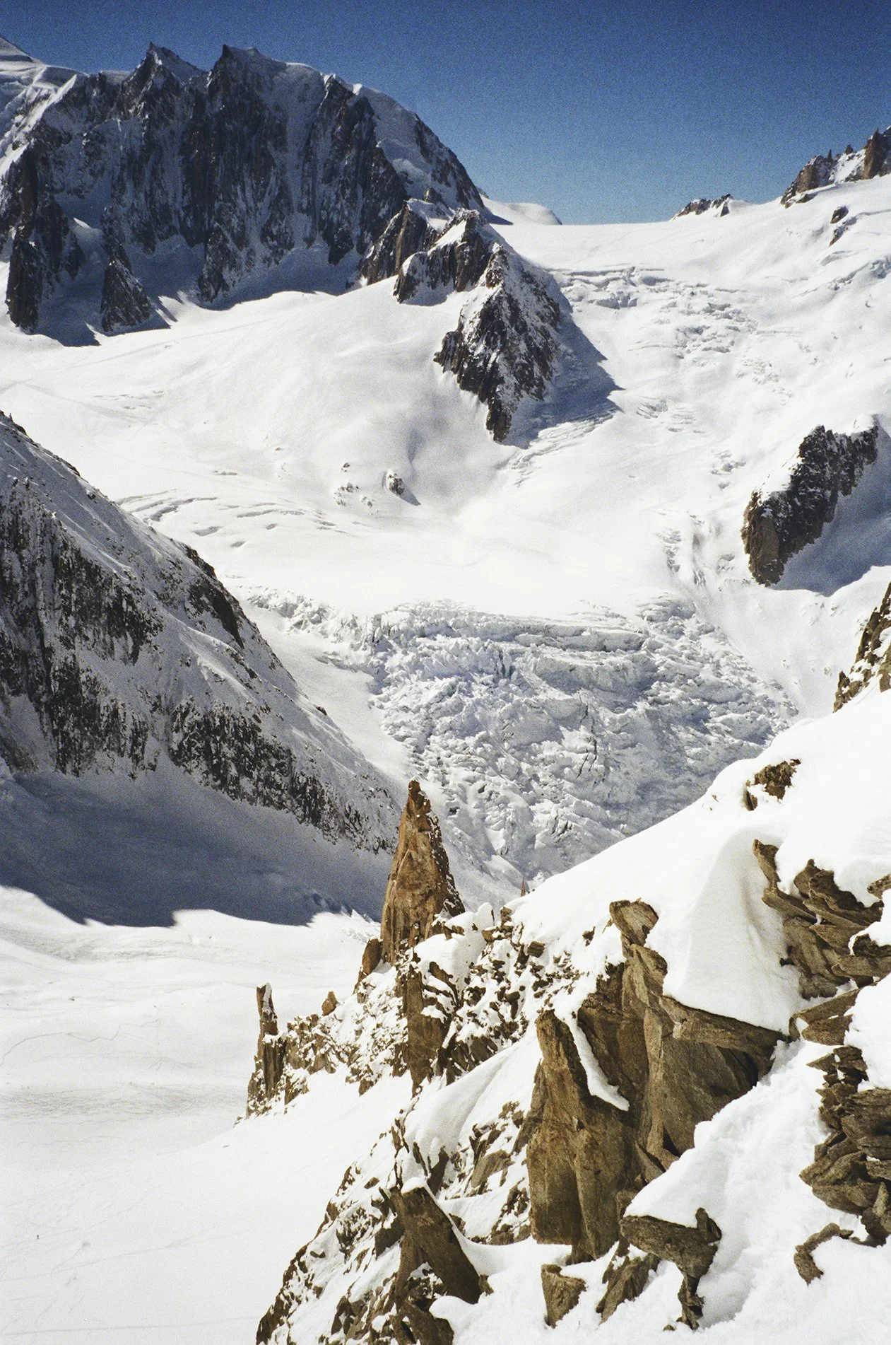 Snow-covered mountain landscape with rugged rocky terrain and alpine peaks under a clear blue sky.