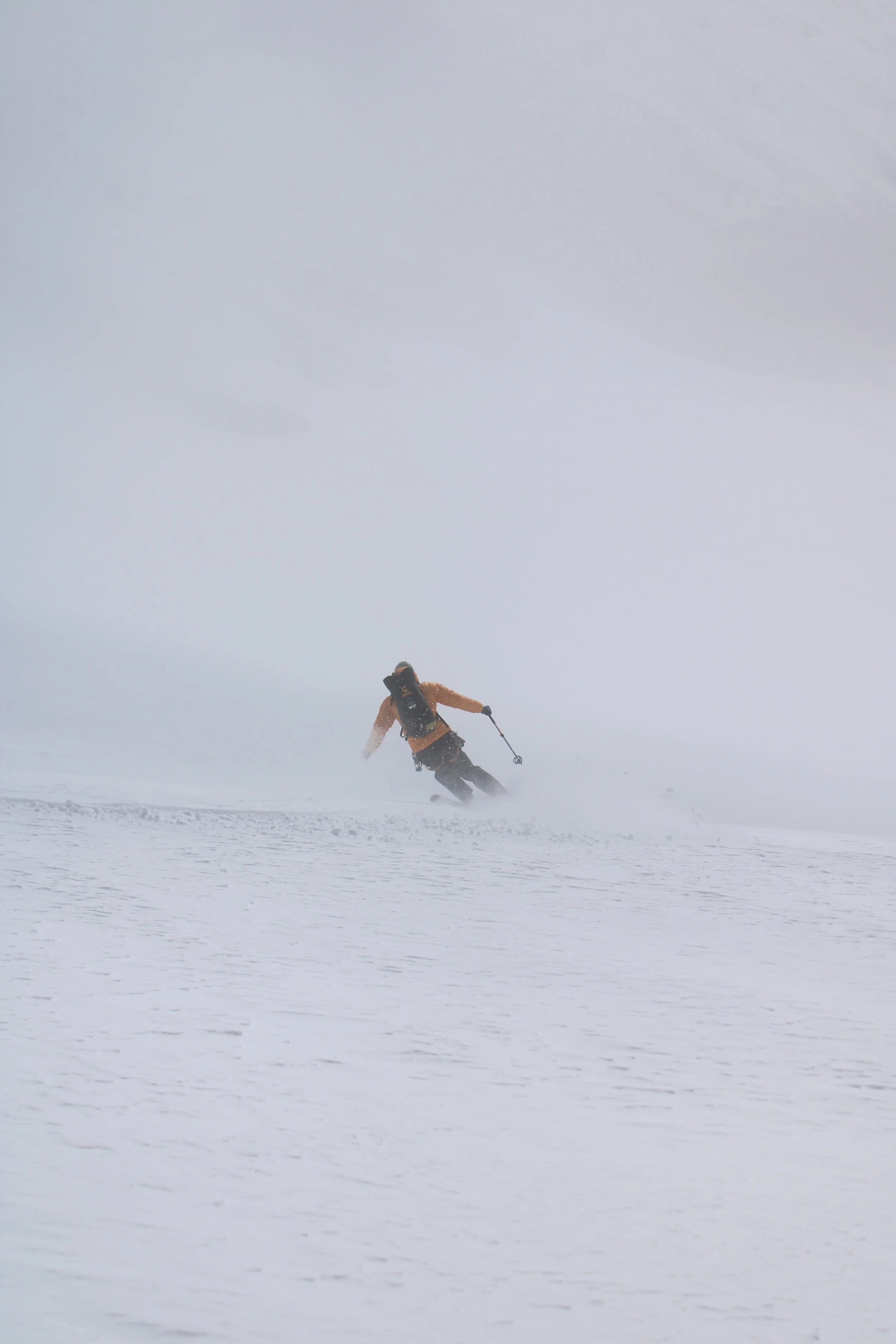 Person skiing on snow-covered terrain in winter, wearing an orange jacket and black pants, holding ski poles, with a cloudy sky above.