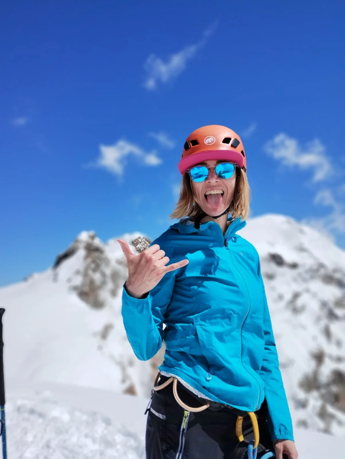 Woman in blue outdoor jacket, wearing a pink helmet and reflective sunglasses, making a shaka sign with her hand, standing on snowy mountain with peaks and a blue sky in the background.